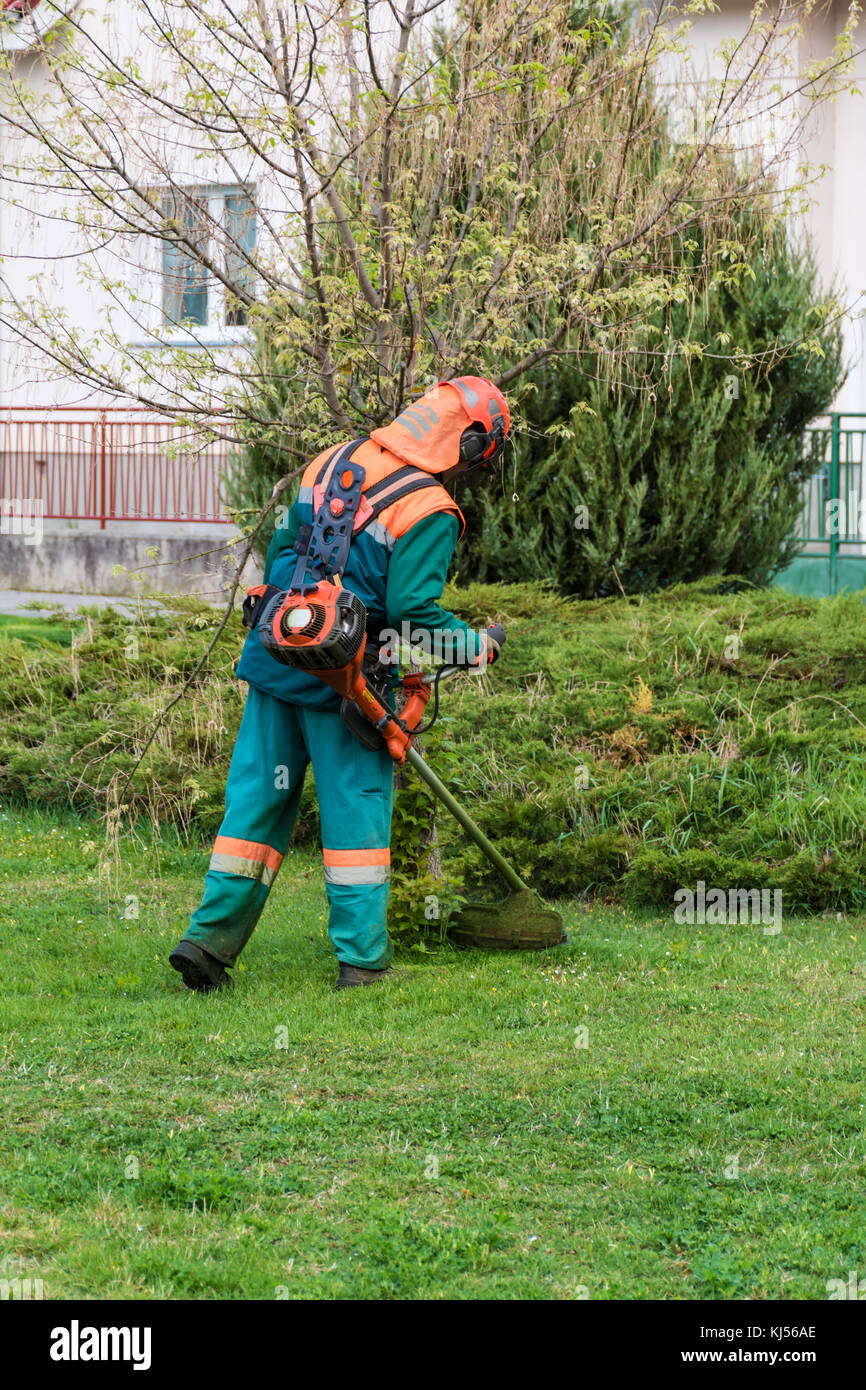 Man in overall and safety helmet trims overgrown grass by grass cutter Stock Photo