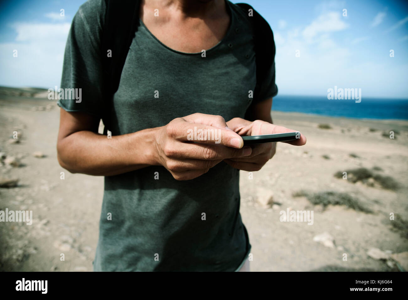 closeup of a young caucasian man carrying a backpack using a smartphone in a natural landscape, with the ocean in the background, with a vignette adde Stock Photo