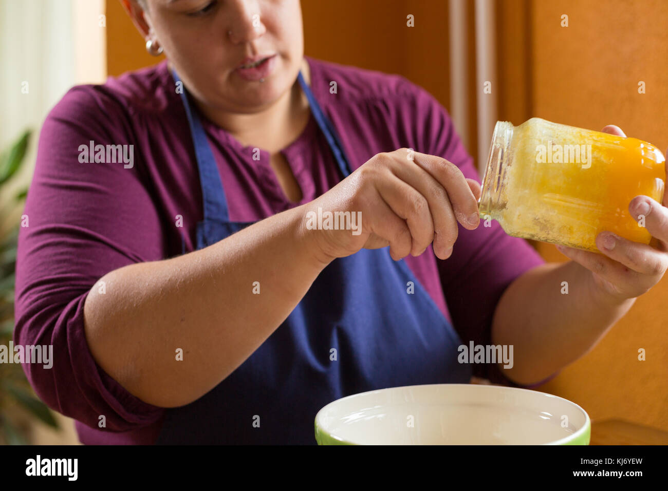 Close up photo of young female holding a jar of organic honey above a green bowl with one hand, getting spoon of honey from the jar Stock Photo