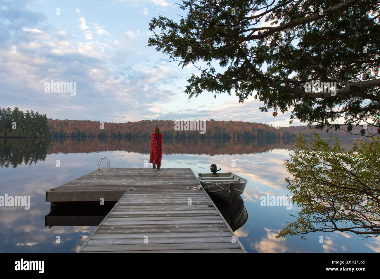 Woman wrapped in plaid blanket on dock looking at fall colors reflected in a lakes surface at sunrise Stock Photo
