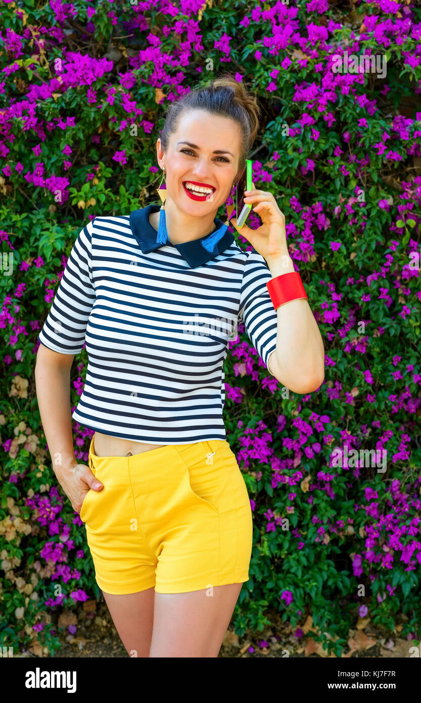 Colorful Freshness. happy young woman in yellow shorts and stripy shirt near colorful magenta flowers bed talking on a mobile phone Stock Photo