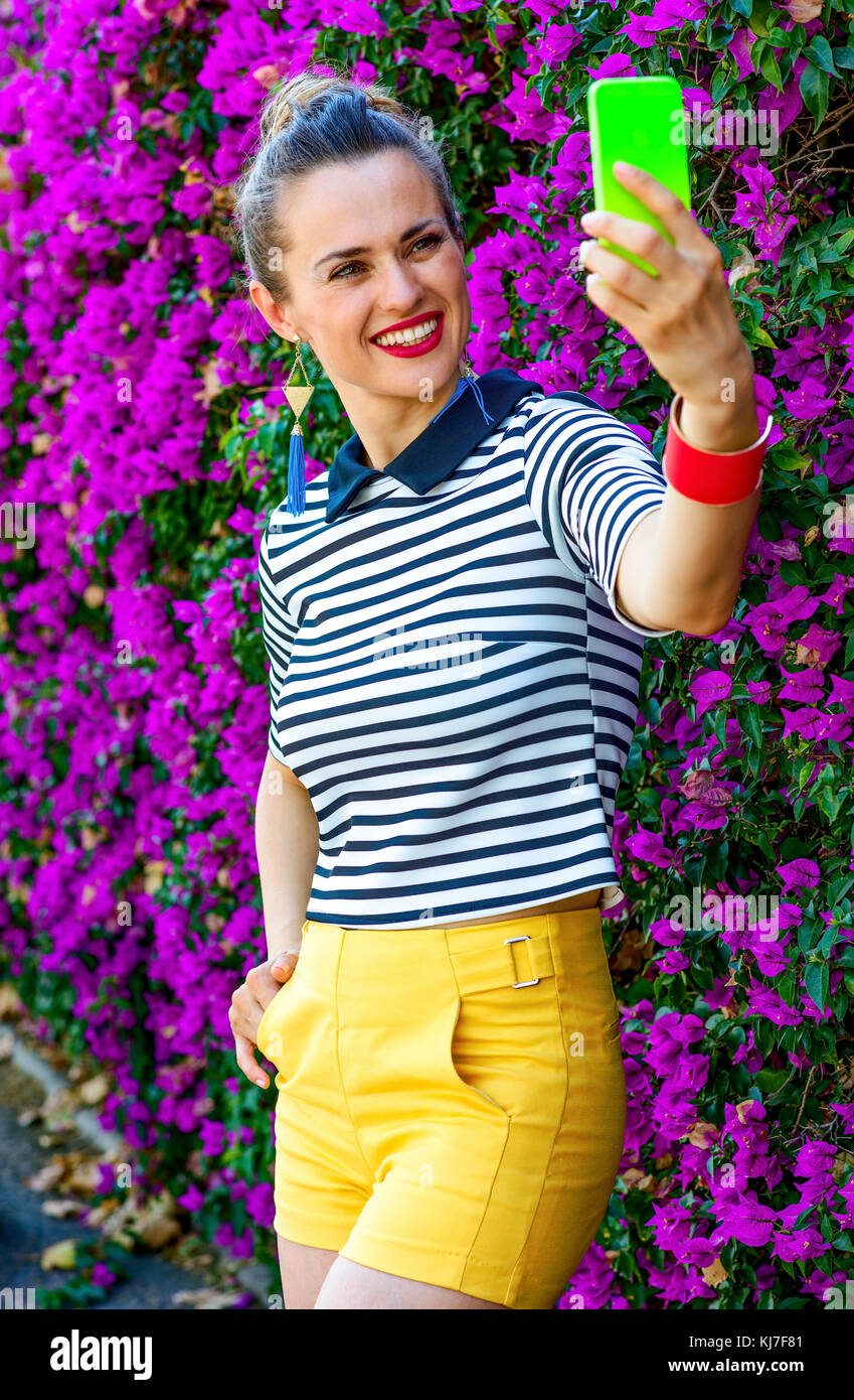 Colorful Freshness. happy modern woman in yellow shorts and stripy shirt in the front of colorful magenta flowers bed with smartphone taking selfie Stock Photo