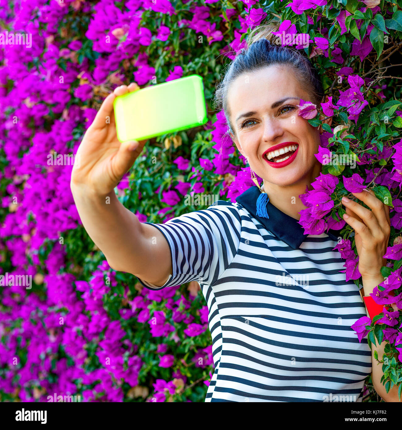 Colorful Freshness. smiling stylish woman in yellow shorts and stripy shirt near colorful magenta flowers bed with phone taking selfie Stock Photo
