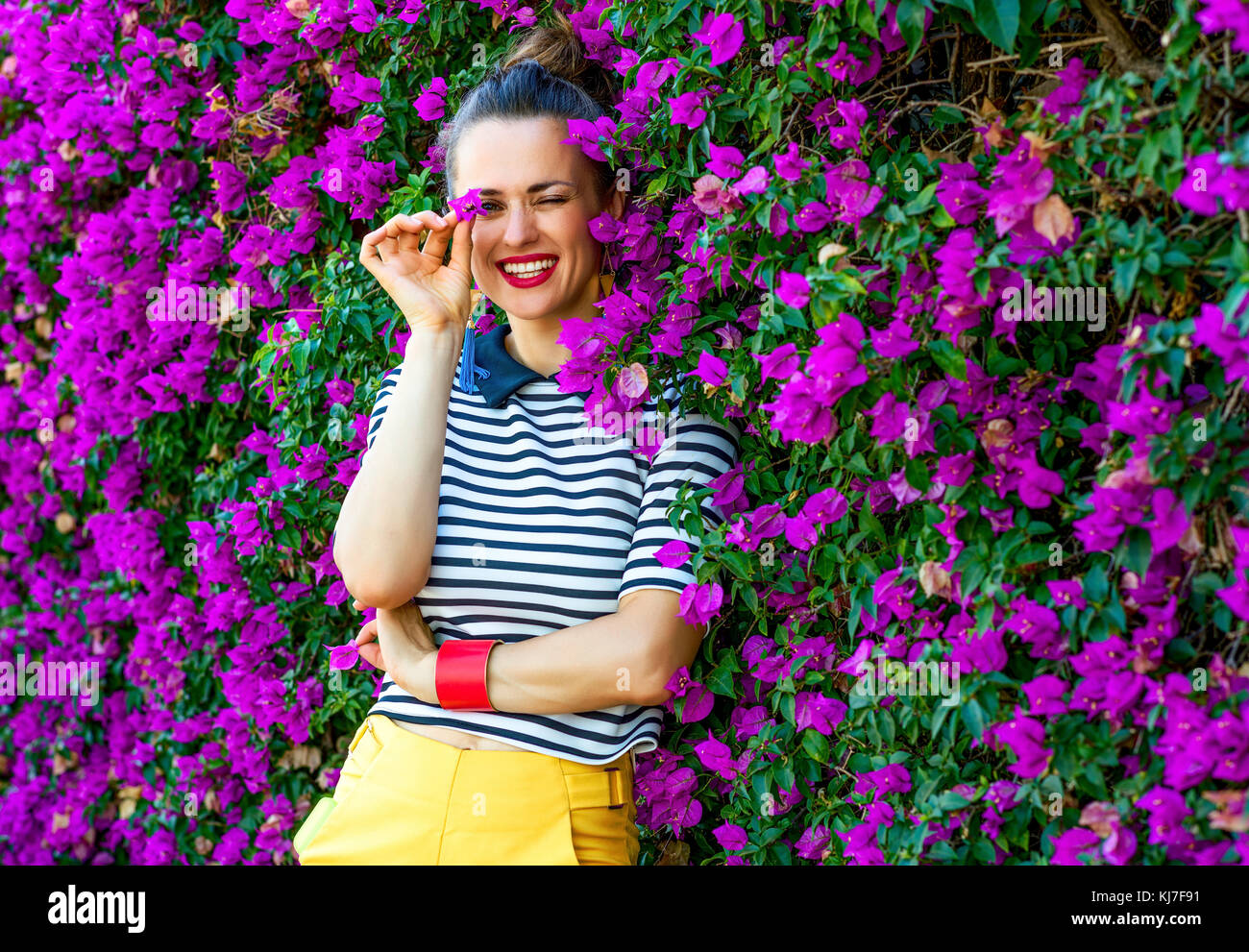 Colorful Freshness. happy stylish woman in yellow shorts and stripy shirt near colorful magenta flowers bed having fun time Stock Photo