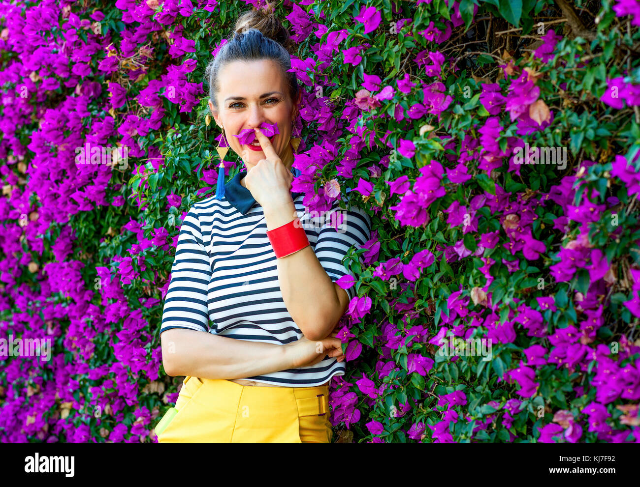 Colorful Freshness. smiling trendy woman in yellow shorts and stripy shirt in the front of colorful magenta flowers bed having fun time Stock Photo