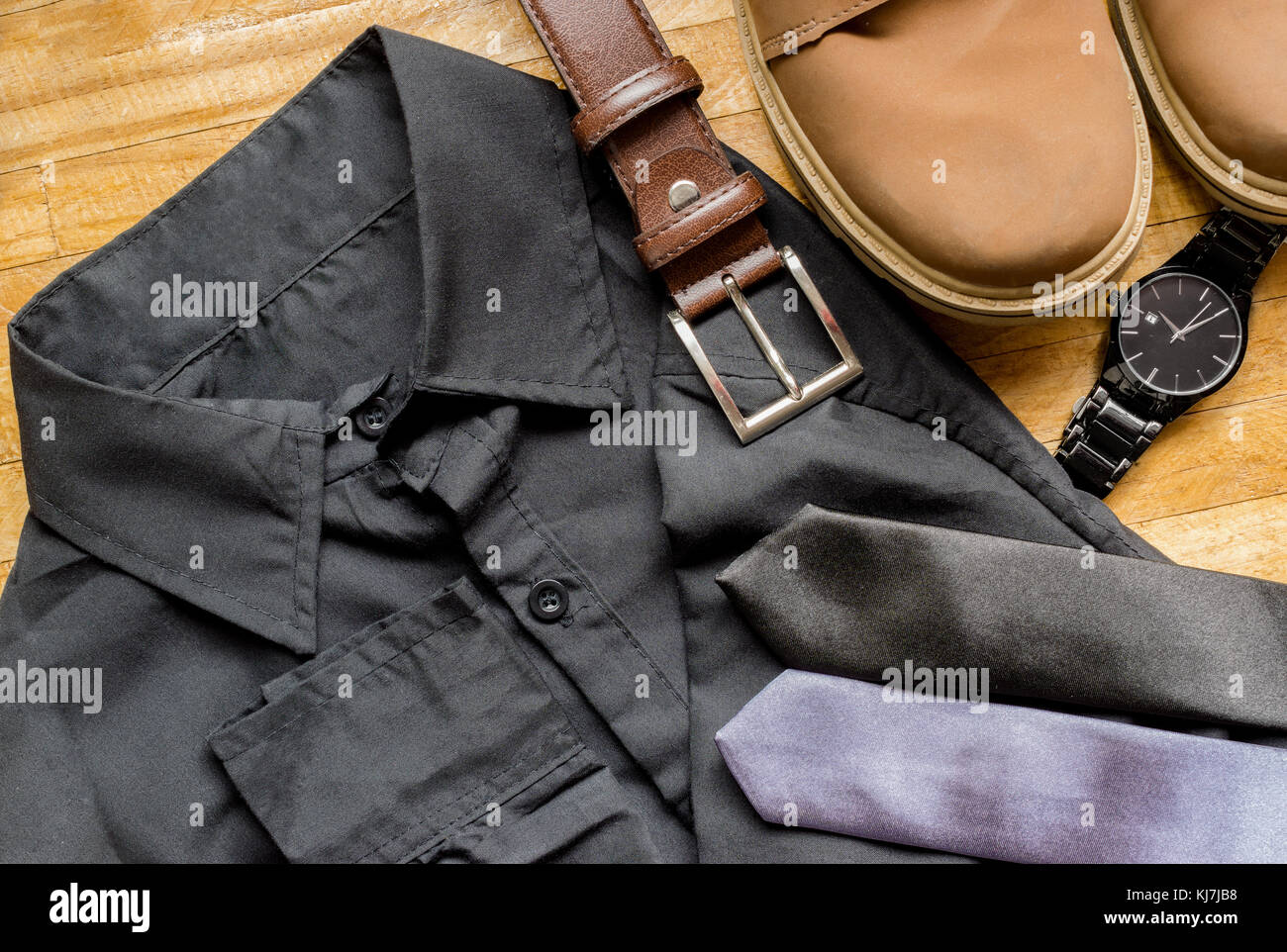 Men's clothes on top of a wooden surface, showing a black dress shirt, brown leather belt, brown boots, neckties and black wrist watch Stock Photo