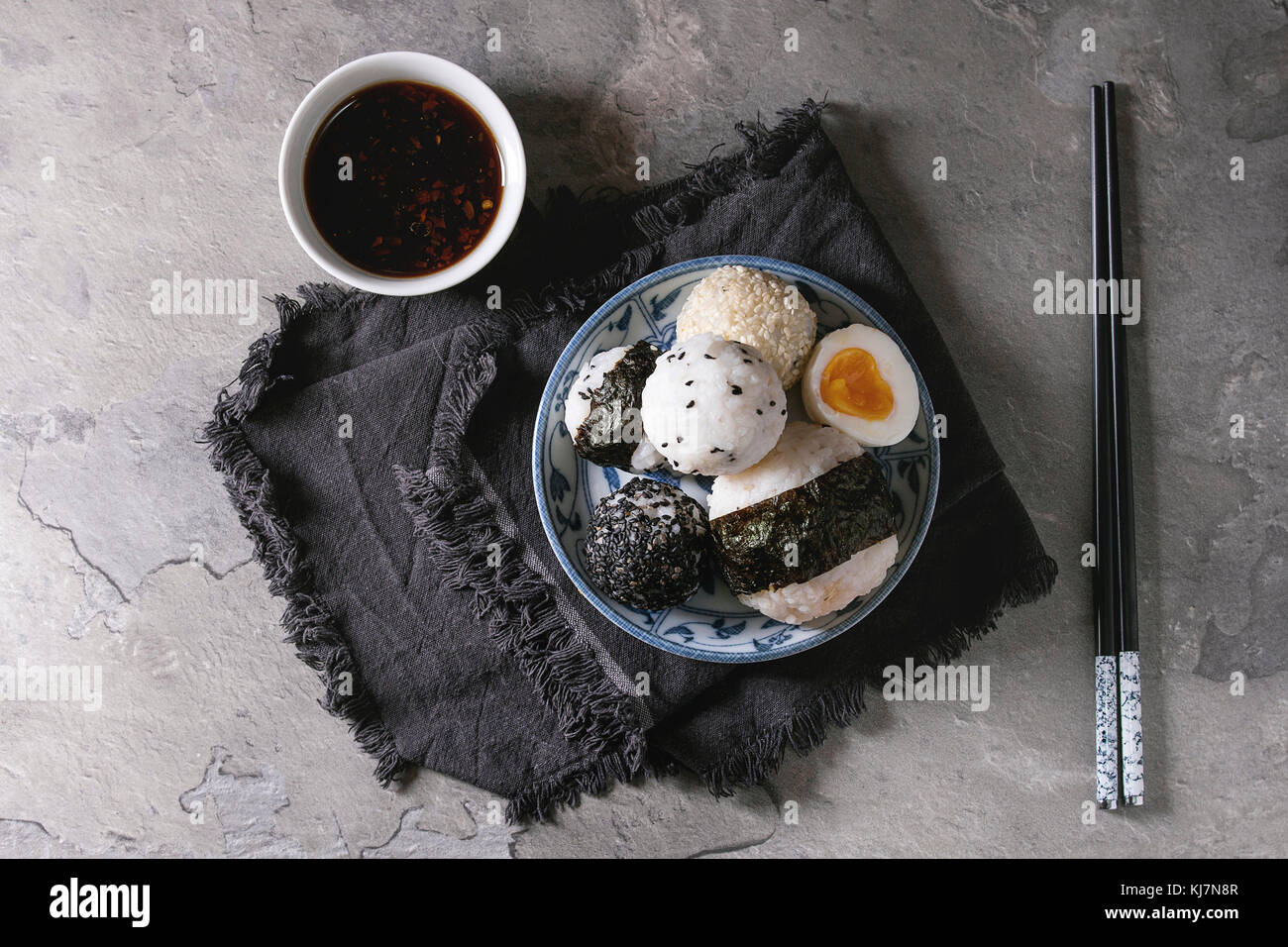 Blue plate with different size rice balls with black sesame and seaweed nori, served with soft boiled eggs, soy sauce, chopsticks over gray table. Asi Stock Photo