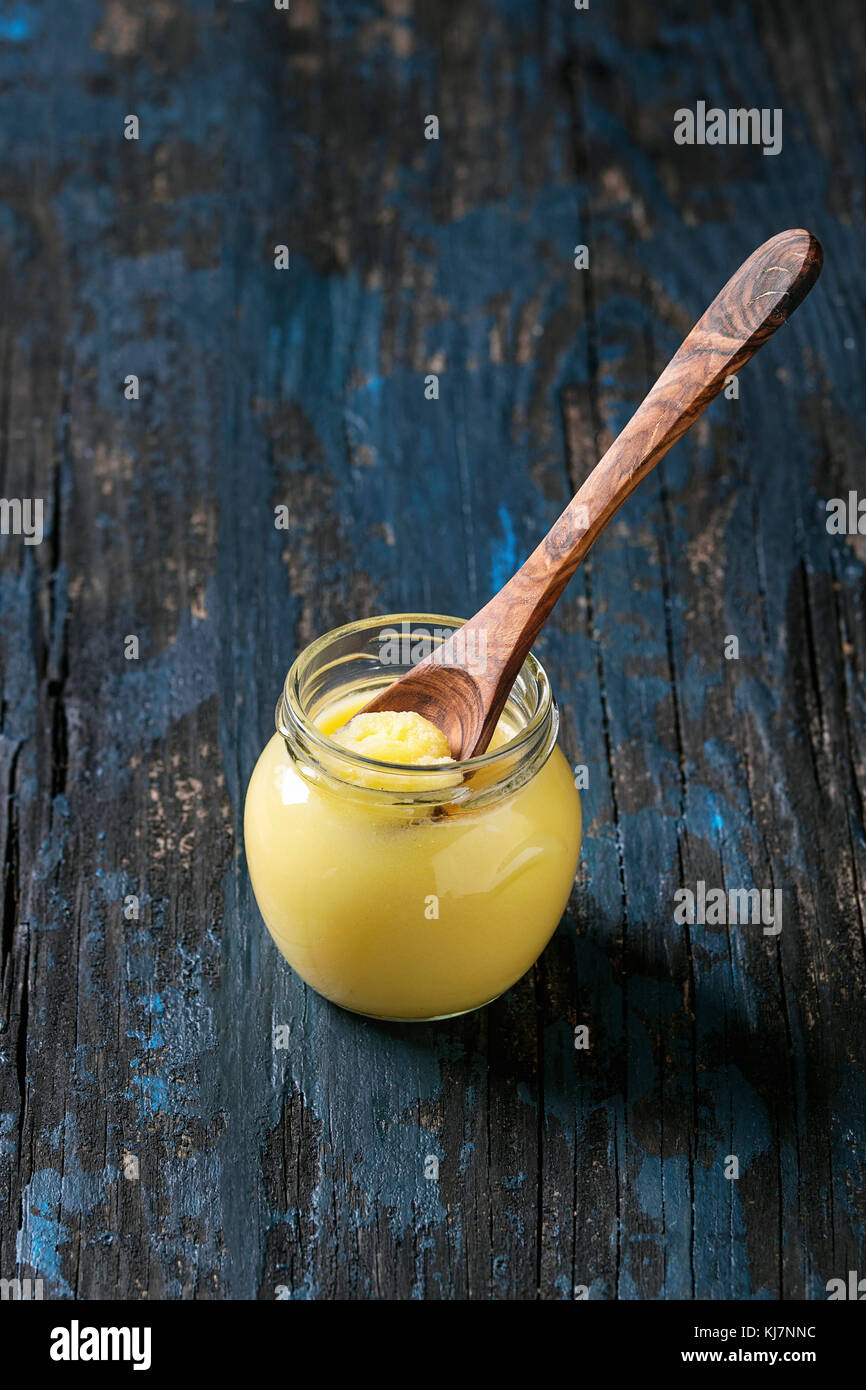 Glass of drawn ghee butter standing with wood spoon over old dark blue