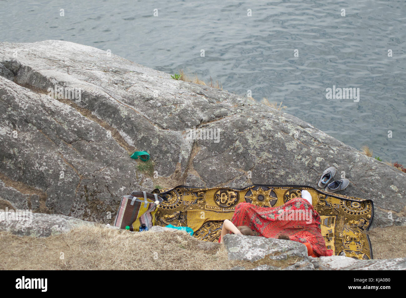 Looking down at partial view of 2 unrecognizable people seated on a colorful patterned blanket on a rock enjoying view of the water below. Stock Photo