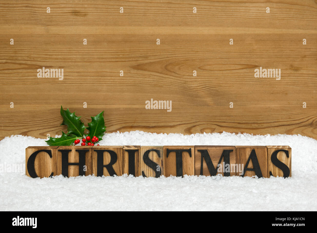 The word CHRISTMAS made from wooden block letters on some snow with a sprig of holly and a rustic wooden background with copy space to add your own me Stock Photo