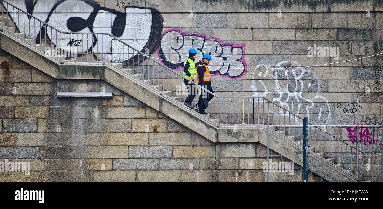 Bremen, Germany - March 8th, 2017 - Two construction workers wearing hi-viz vests and helmets walking down a set of stairs in front of an old graffiti Stock Photo