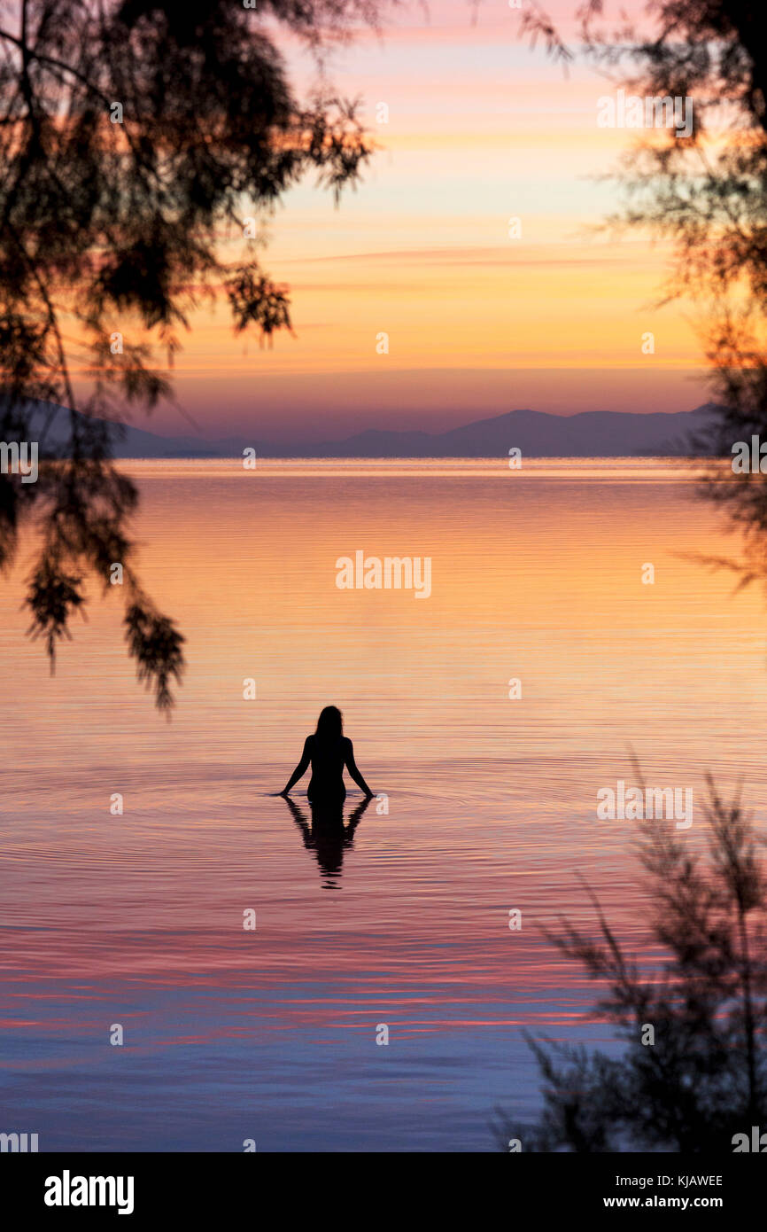 A lone woman bather is reflected as she enters the calm water of the Aegean sea. She is silhouetted against the  flaming colors of a dawn sky. Stock Photo