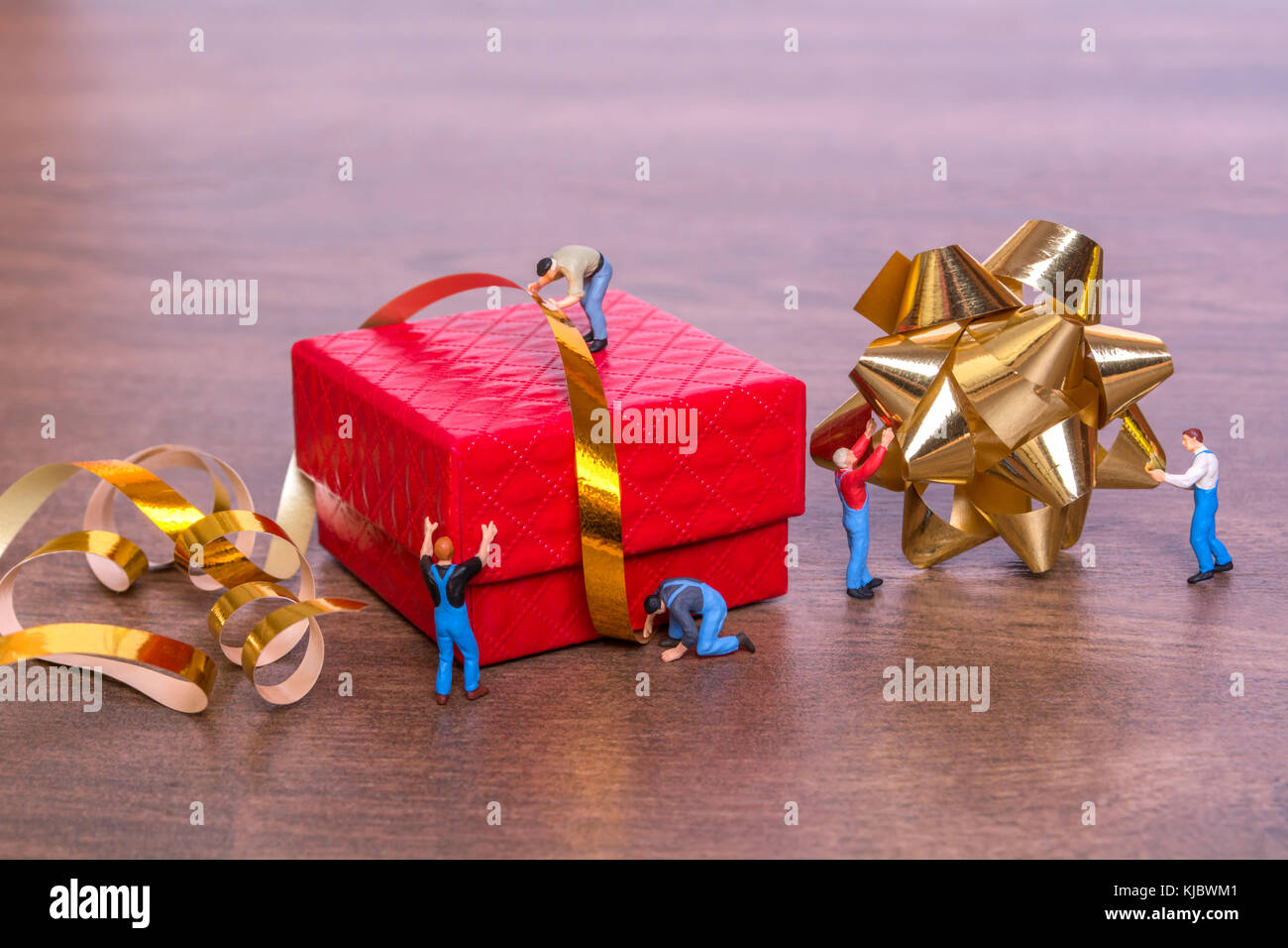 Creative concept with miniature people and a gift box on a wooden background. The process of packing gifts. Gold ribbon, bow, festive beads. Creative  Stock Photo