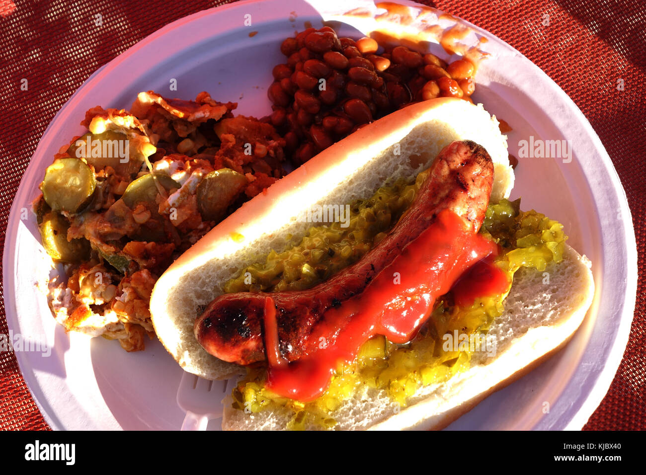 A bbq'd brat covered with ketchup, mustard and relish is plated on a paper plate along with baked beans and a Mexican pasta salad. Stock Photo