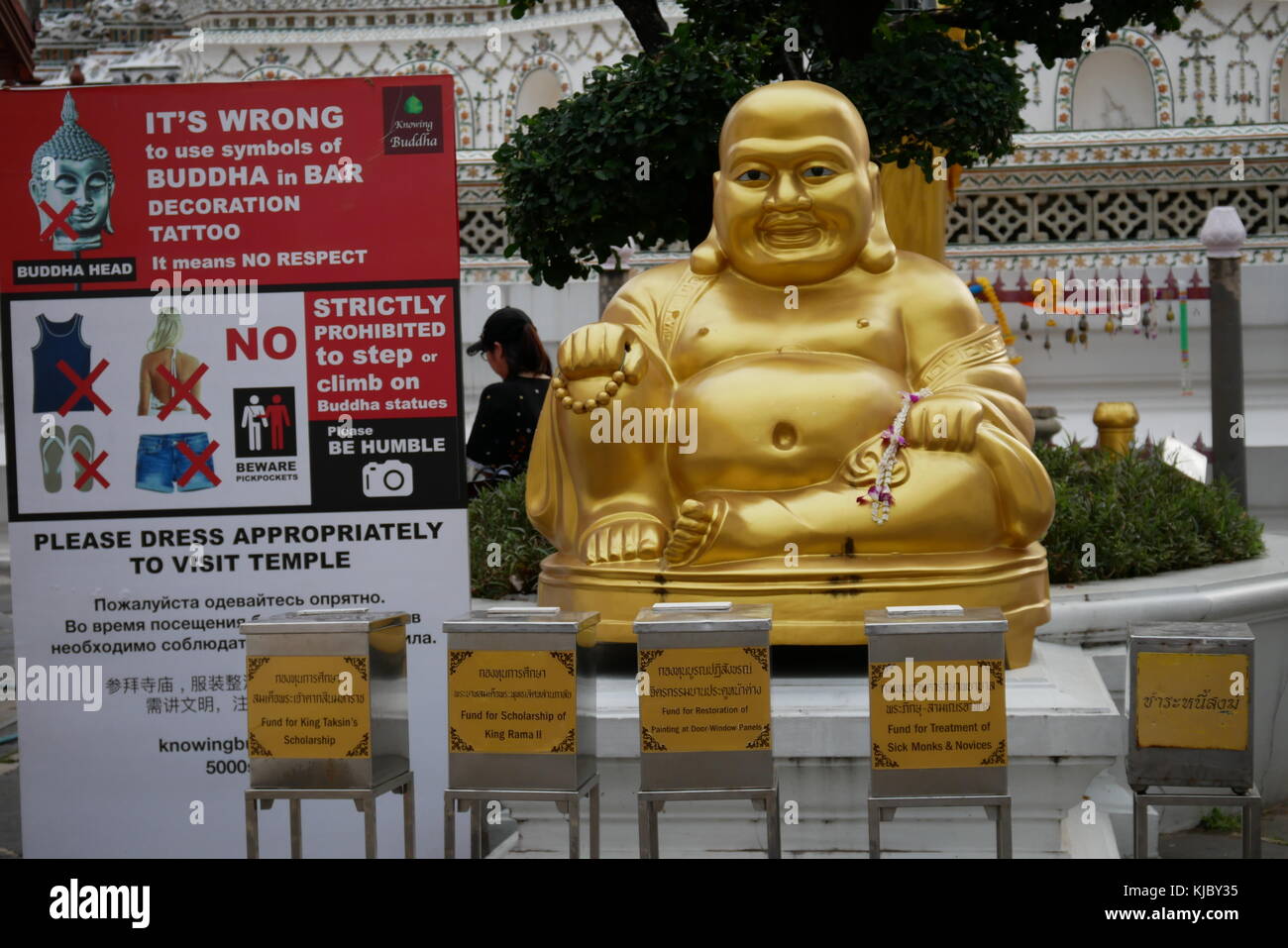 A Laughing Buddha golden statue sits in front of Wat Arun and next to a official banner forbidding the use of Buddha as a decoration or tattoo design. Stock Photo