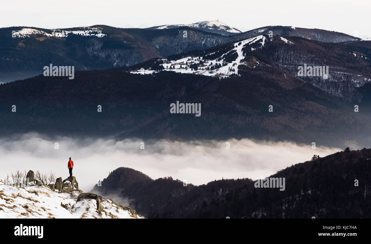 A hiker standing on a snowy peak looking at a beautiful and dramatic cloud inversion. Wanderlust in the Vosges mountains in winter, France. Stock Photo