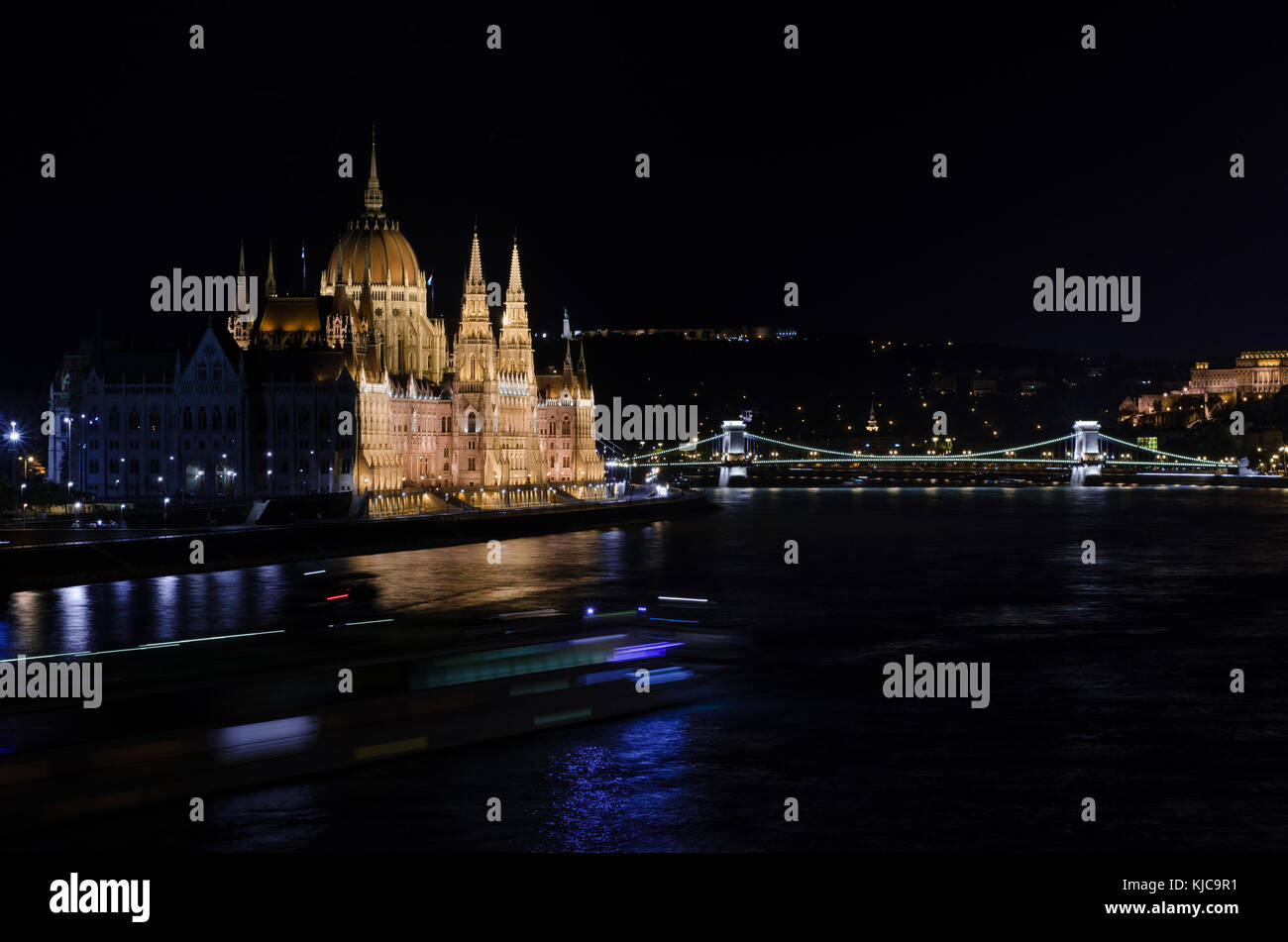 The Hungarian Parliament, Országház, and the Széchenyi Lánchíd bridge lit at night and looking over the Danube river, with a barge passing. Stock Photo