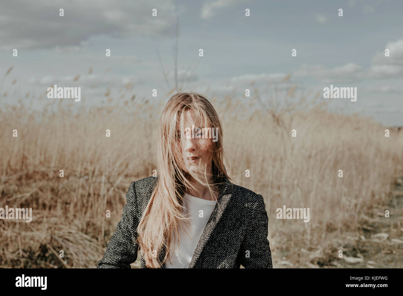 Pretty and young european woman with face covered  by her blonde hair looking at camera. Girl posing outdoor on bulrush background. Close shot. Stock Photo
