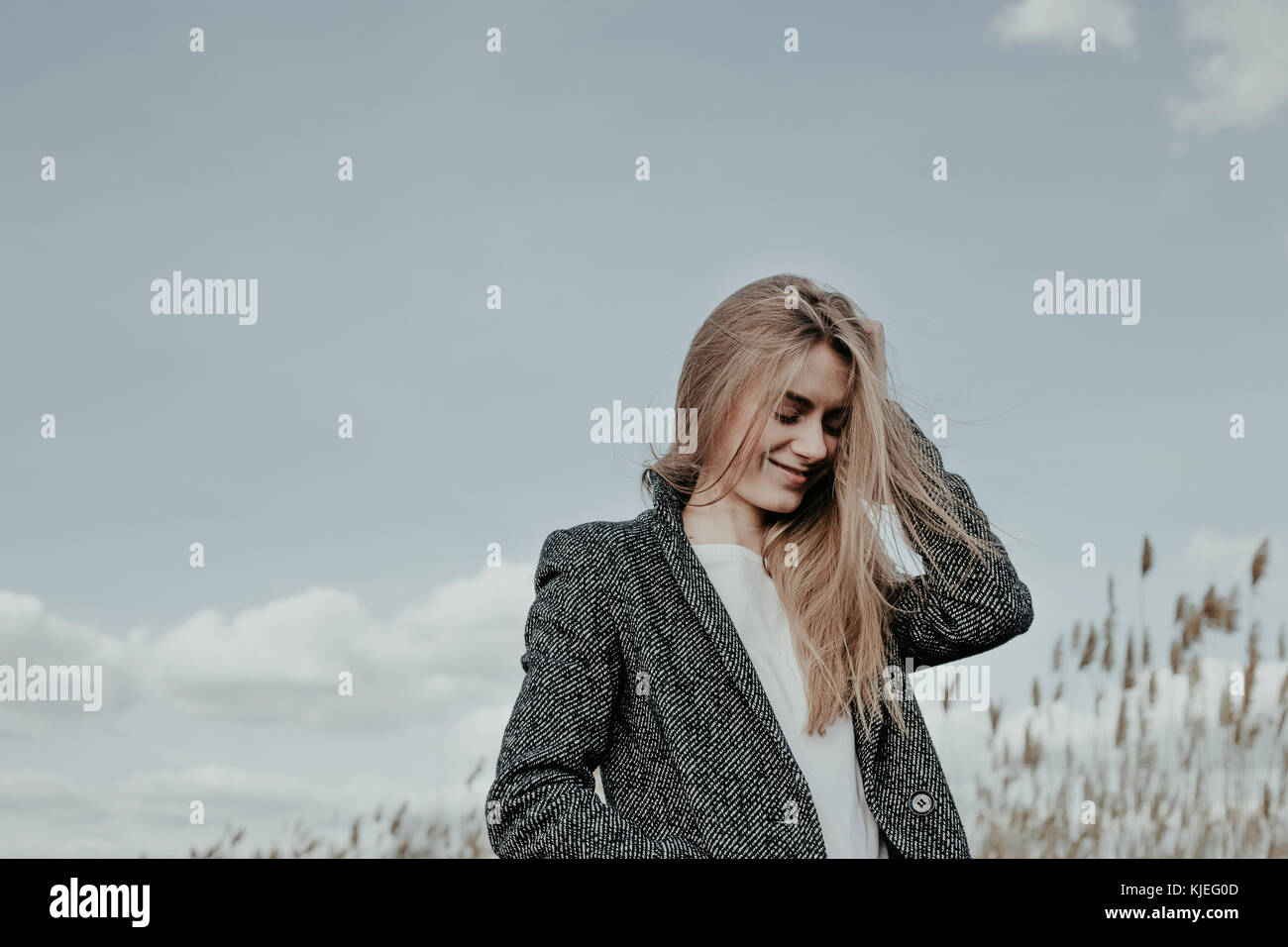 Pretty young and slim woman with long blonde hair posing at camera outdoor on sky background. Cute girl smiling and looking down. Copy space. Stock Photo