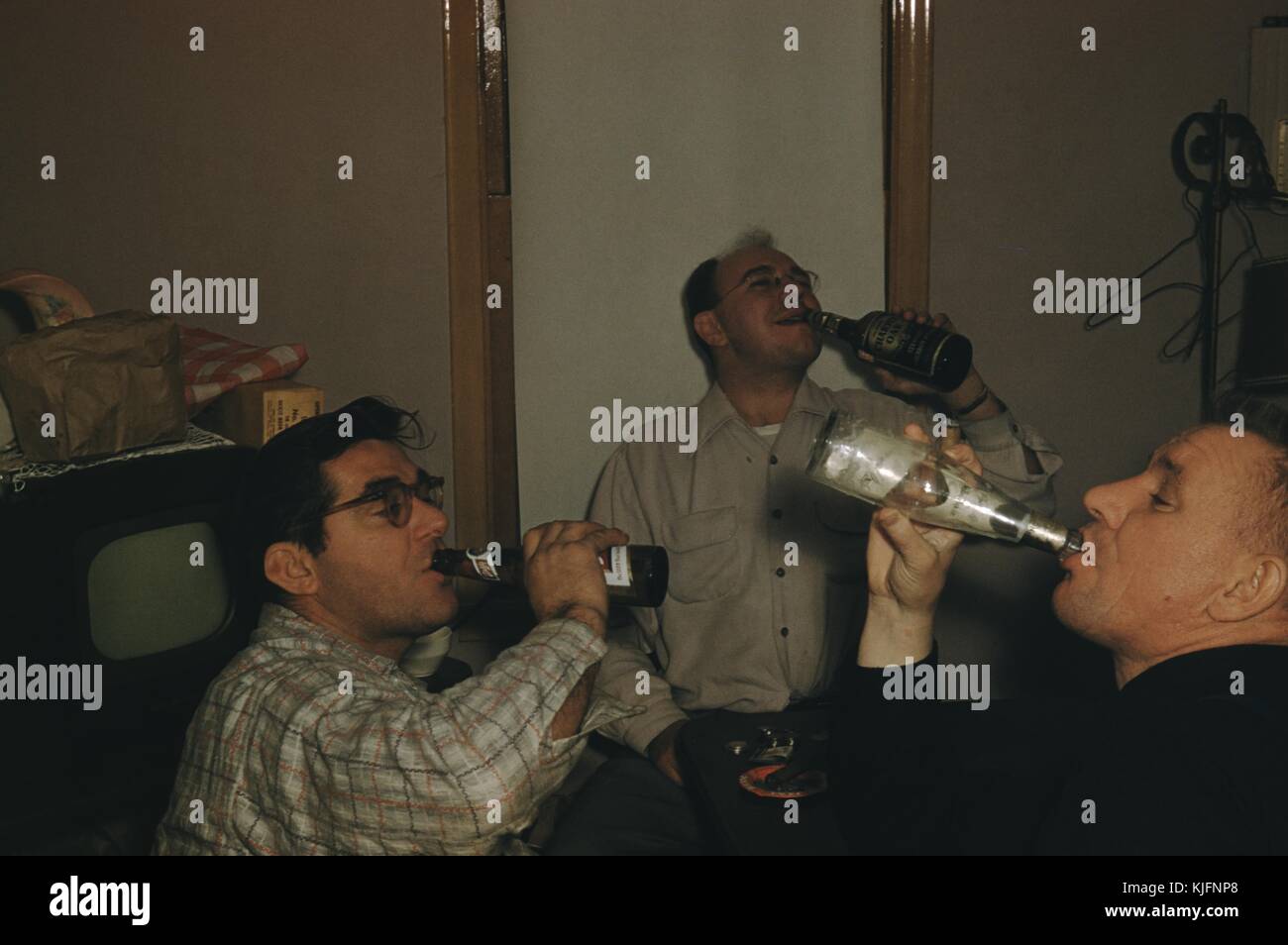 A photograph of three men posing while drinking bottles of beer, they are sitting around a small table where a cigar can be seen resting in an ashtray, there is a small antique television in the background with items piled on top of it, 1952. Stock Photo