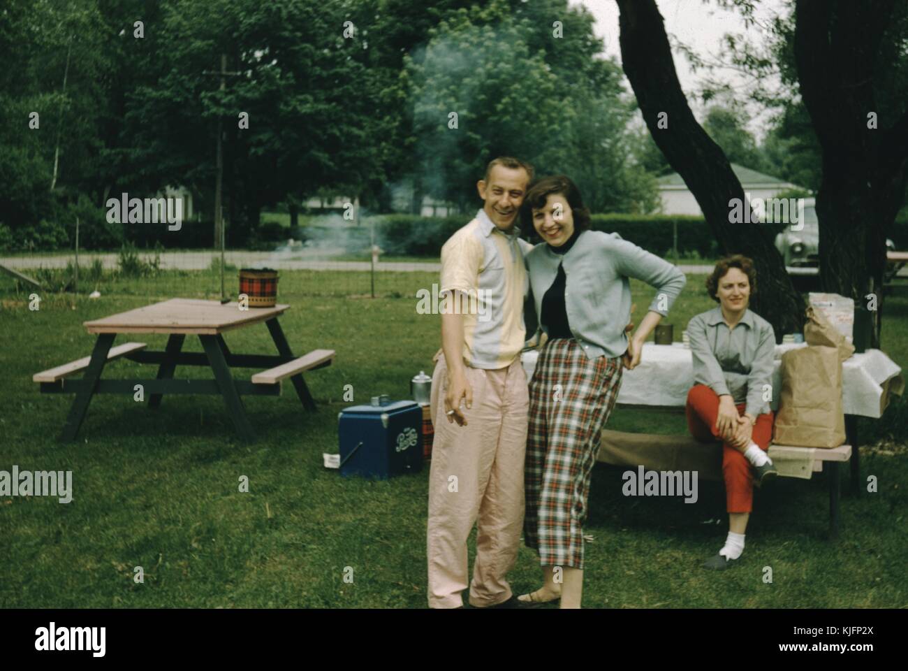 Couple posing for a picture at a park, man is smoking a cigarette, behind them a woman sitting at a picnic table covered with a table cloth, a cooler, and what appear to be bags of groceries, the people all seem to be in 1950s attire, 1952. Stock Photo