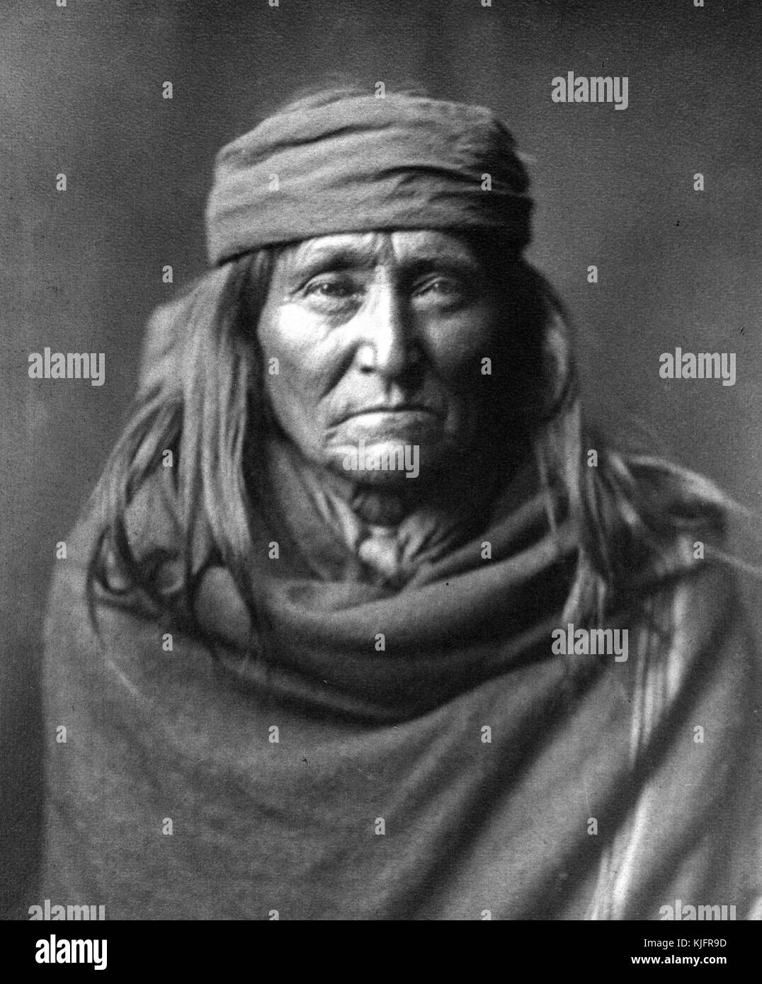Portrait of a mature Native American man with long hair, a head band, and wrapped in a blanket, titled 'Eskay De, A typical, shrewd, old Apache', by Edward S Curtis, 1903. From Portrait of a mature Native American man with long hair, a head band, and wrapped in a blanket, titled 'Eskay De, A typical, shrewd, old Apache', by Edward S Curtis, 1903. From