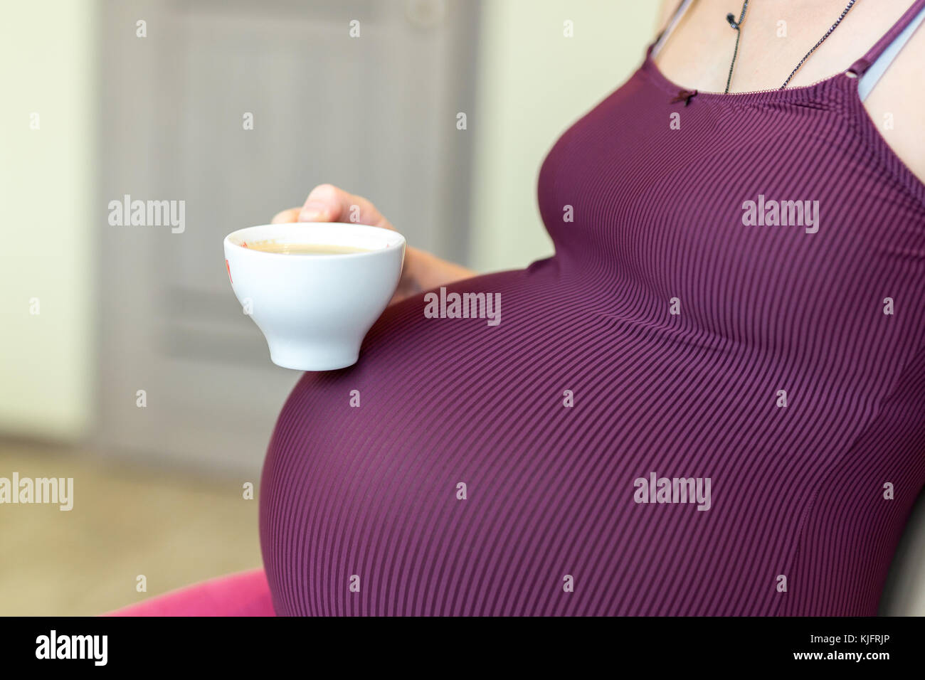 Young pregnant woman sitting in a kitchen and holding a cup od coffee ...