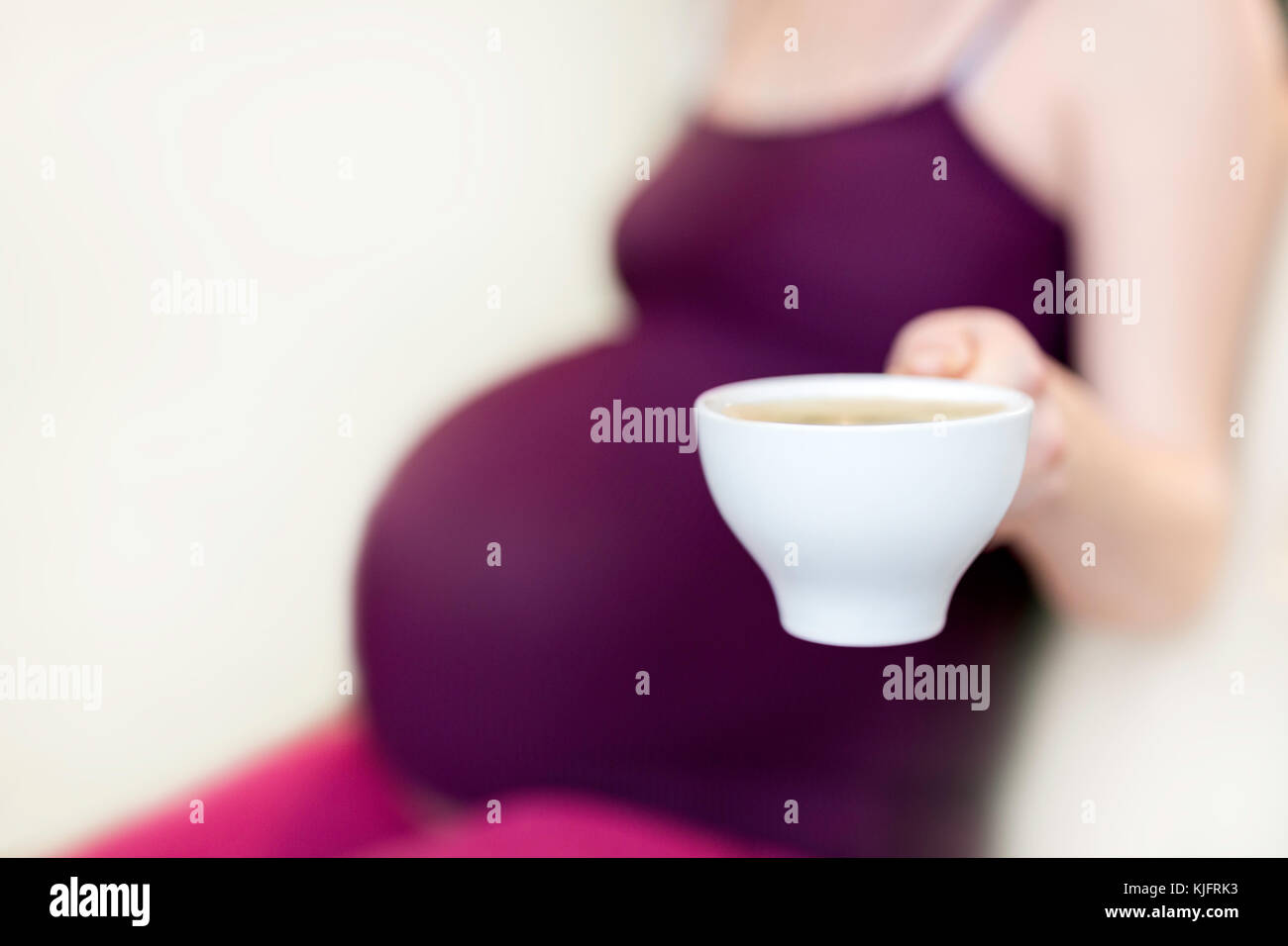 Young pregnant woman sitting in a kitchen and holding a cup od coffee ...