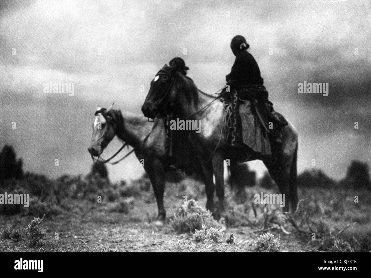 A photograph of two Navajo women on horseback, the woman in the background is obscured by the horse in the foreground, the woman in the foreground wears a dark colored top and a long patterned skirt, they are posed among short desert grass with stormy skies for a backdrop, 1906. From the New York Public Library. Stock Photo