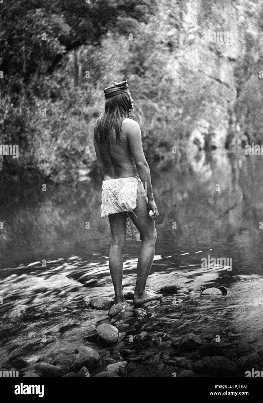 A photograph of a man belonging to the Apache Native American tribe, he is shown standing on rocks with water flowing around him, the man is only wearing a breechcloth and headband, trees and a high cliff face can be seen in the background, 1906. From the New York Public Library. Stock Photo