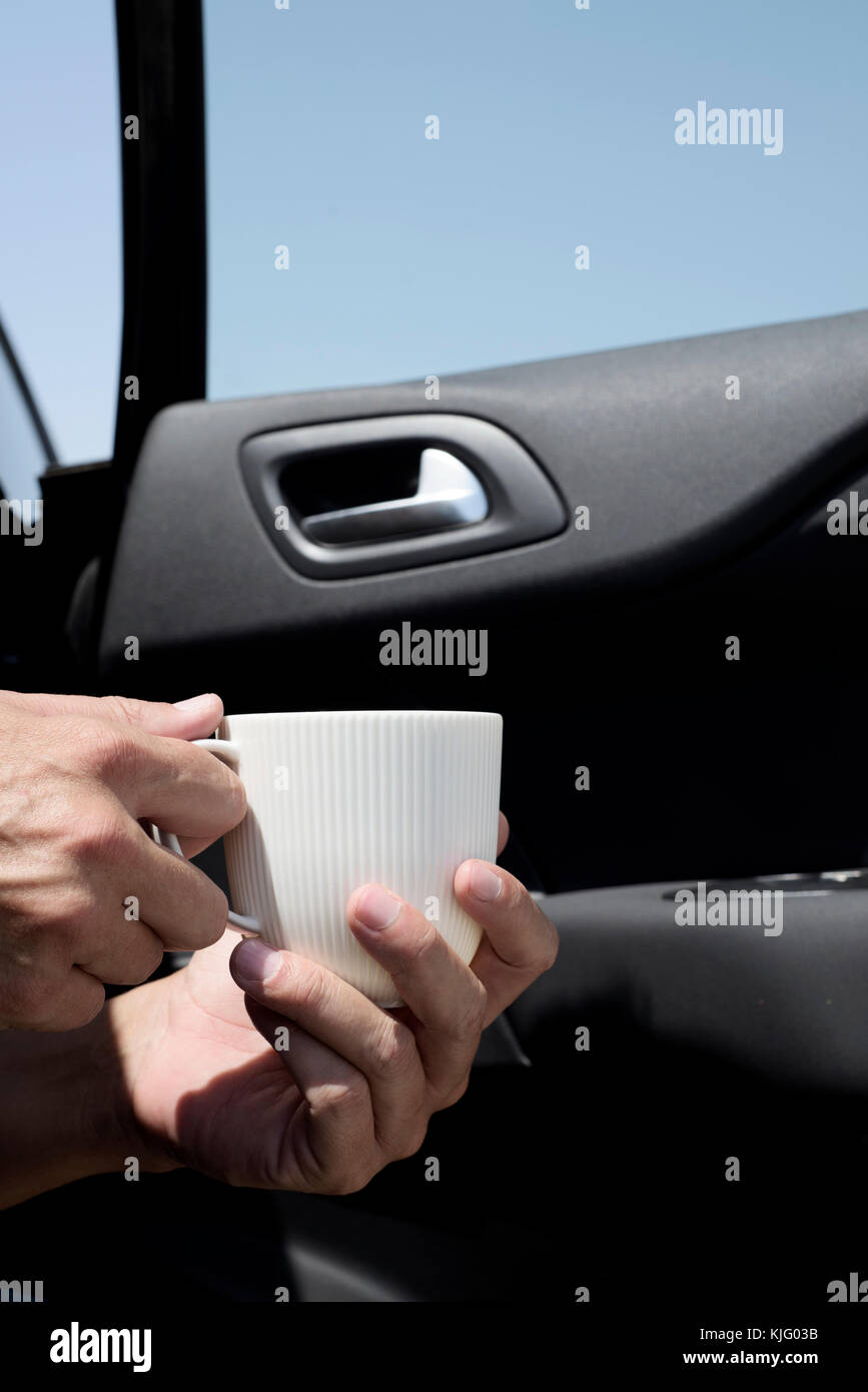 closeup of a young caucasian man with a cup of coffee in his hands sitting at the front passenger seat of a car with its door open Stock Photo