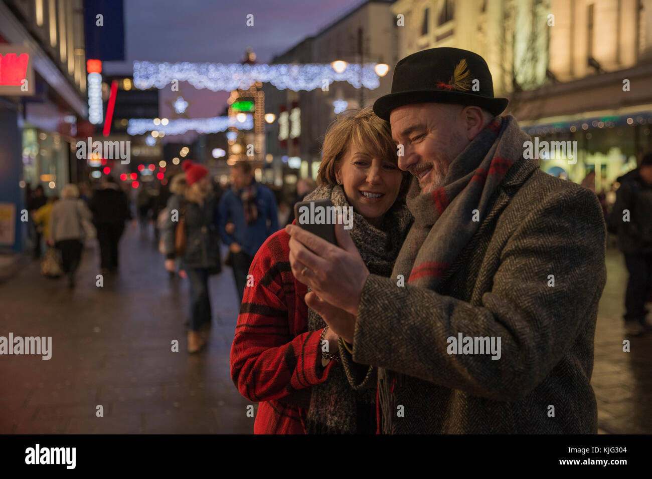 Mature couple are in the city centre on an evening at Christmas time. They are using a smart phone to take a selfie together. Stock Photo