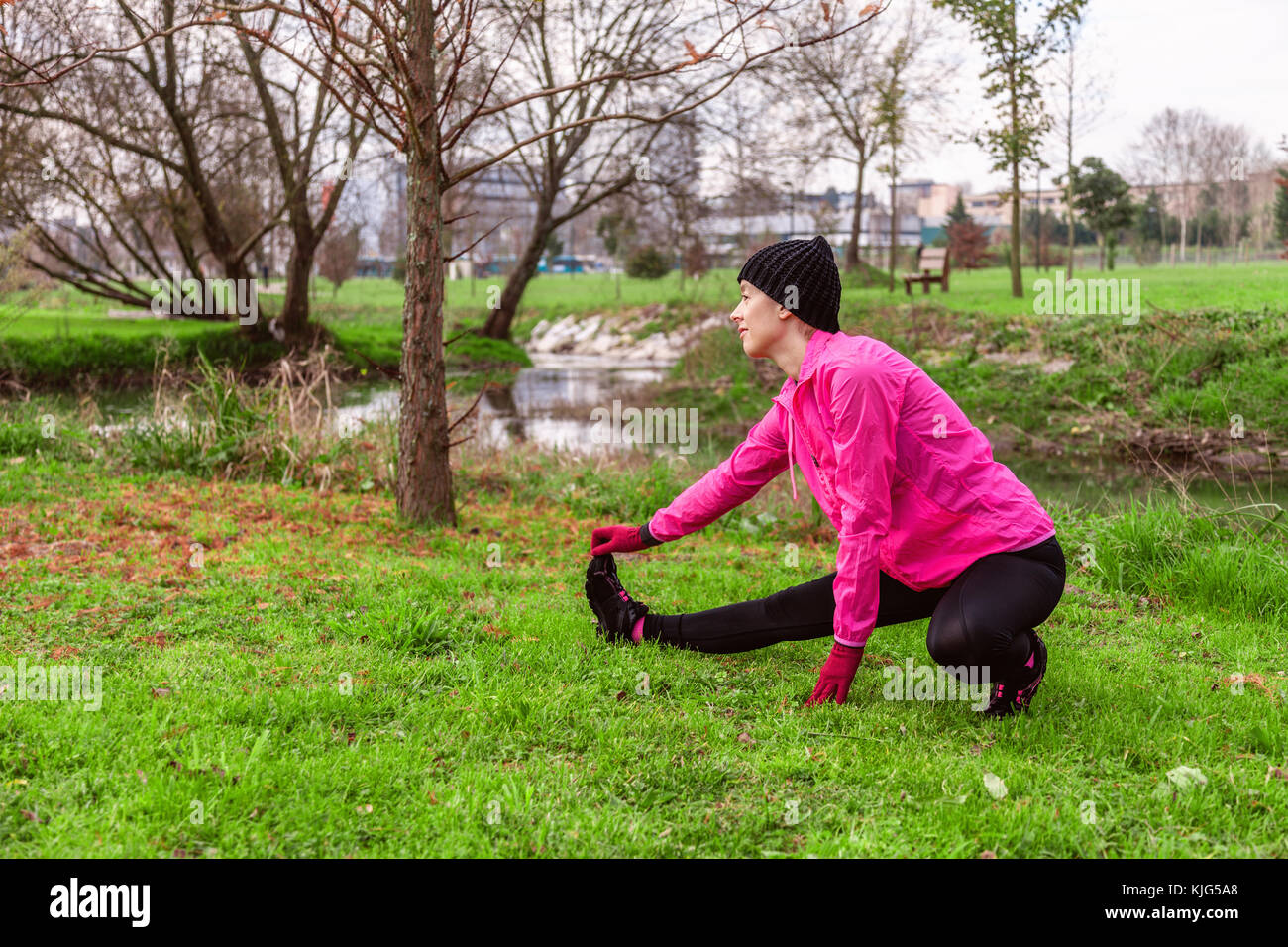 Young woman warming up and stretching the legs before running on a cold winter, autumn of fall day in an urban park. Female athlete wearing pink windb Stock Photo