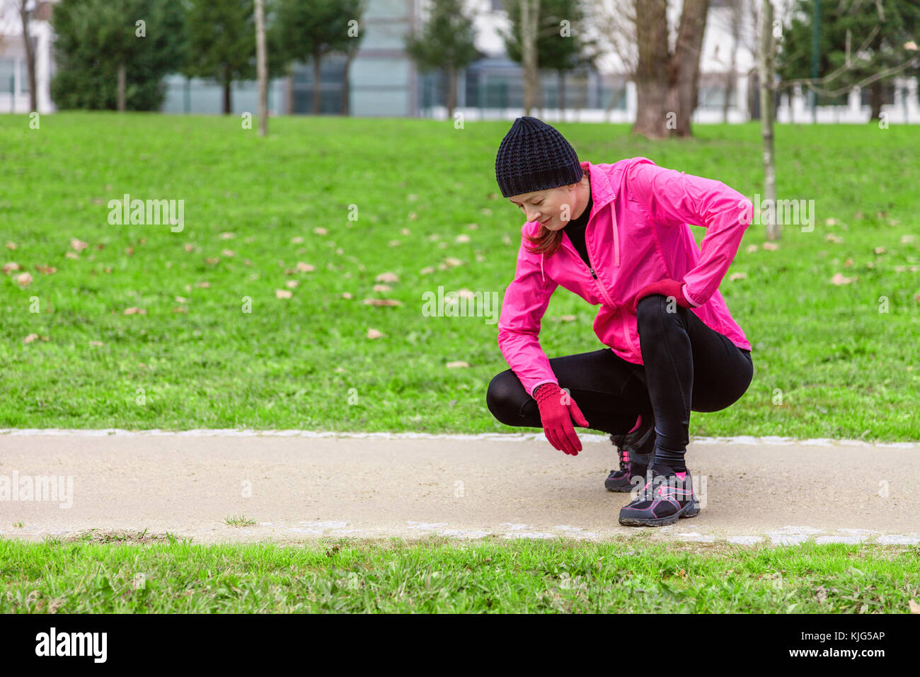 Young woman exhausted after train on a cold winter day on the training track of an urban park. Female athlete wearing pink windbreaker, beanie, gloves Stock Photo