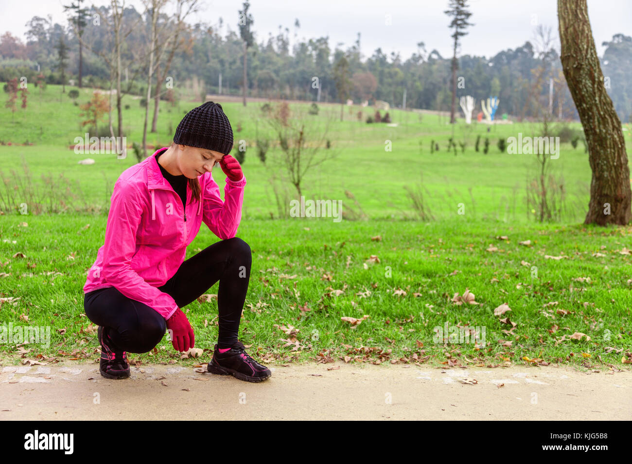 Young woman feeling lightheaded or with headache after train on a cold winter day on the training track of an urban park. Female athlete wearing pink  Stock Photo