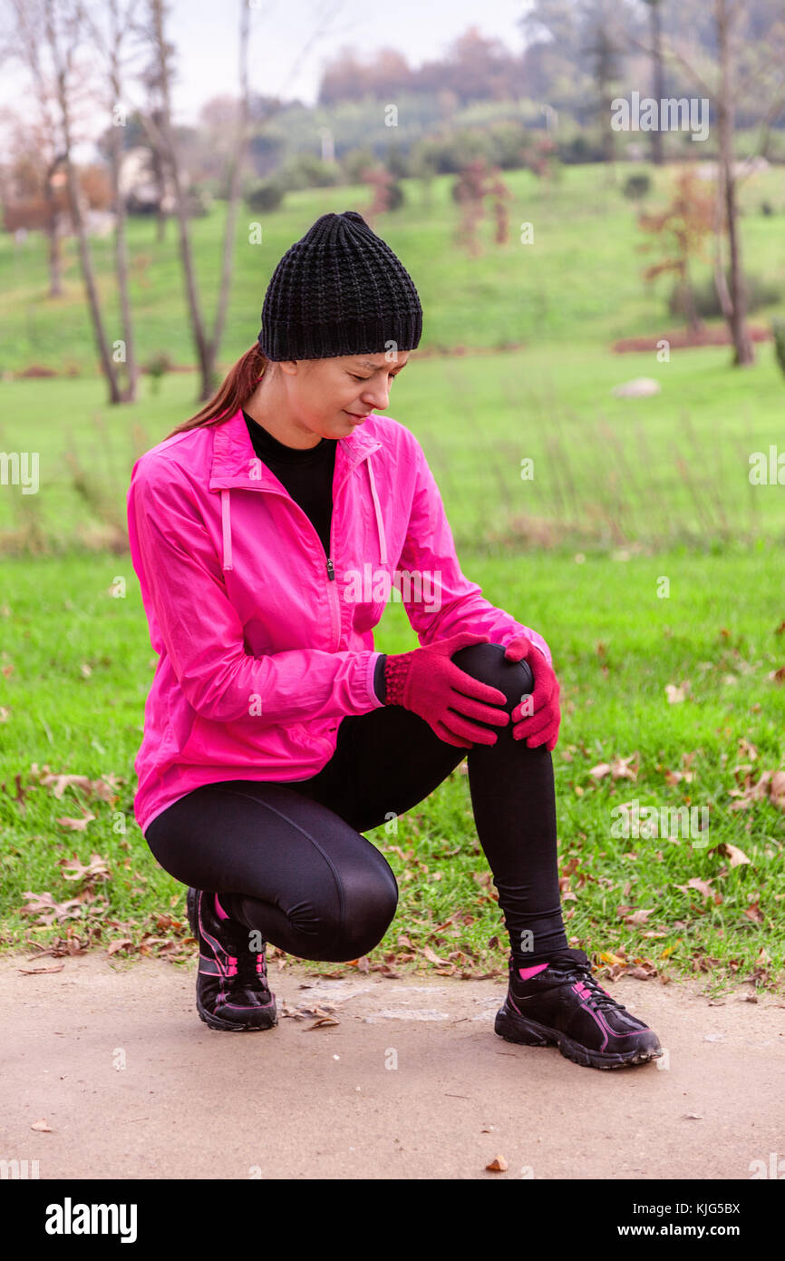Female athlete hurting from a knee injury on a cold winter day on the training track of an urban park. Young woman wearing pink windbreaker, beanie, g Stock Photo