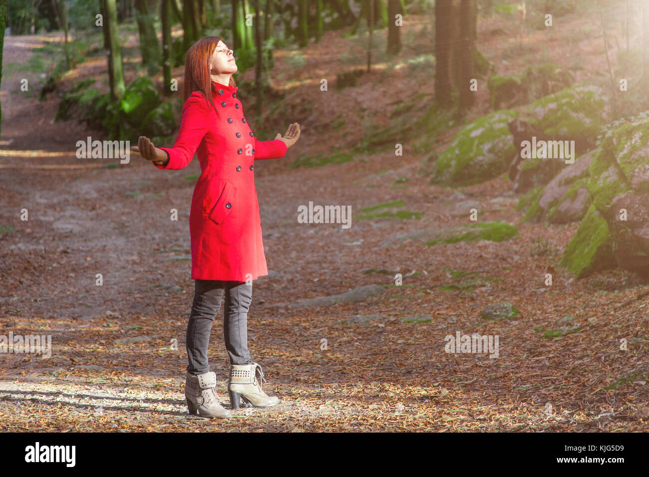 Woman enjoying the warmth of winter sunlight alone on forest park path with arms open receiving rays of light, wearing a red long coat or overcoat Stock Photo