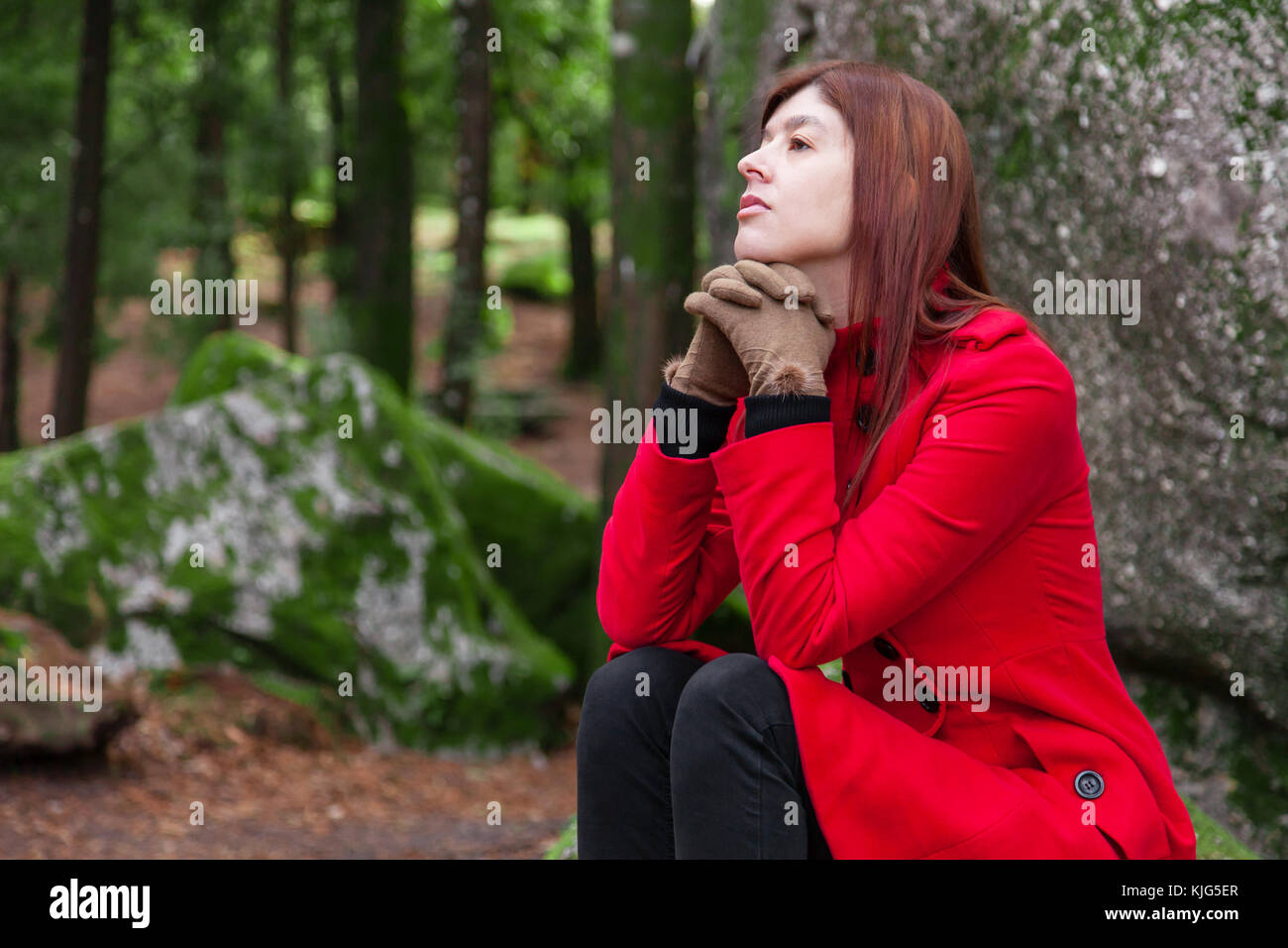 Depressed and sad young woman feeling depression sitting on forest, looking up with melancholic thinking, wearing a red long coat or overcoat during f Stock Photo