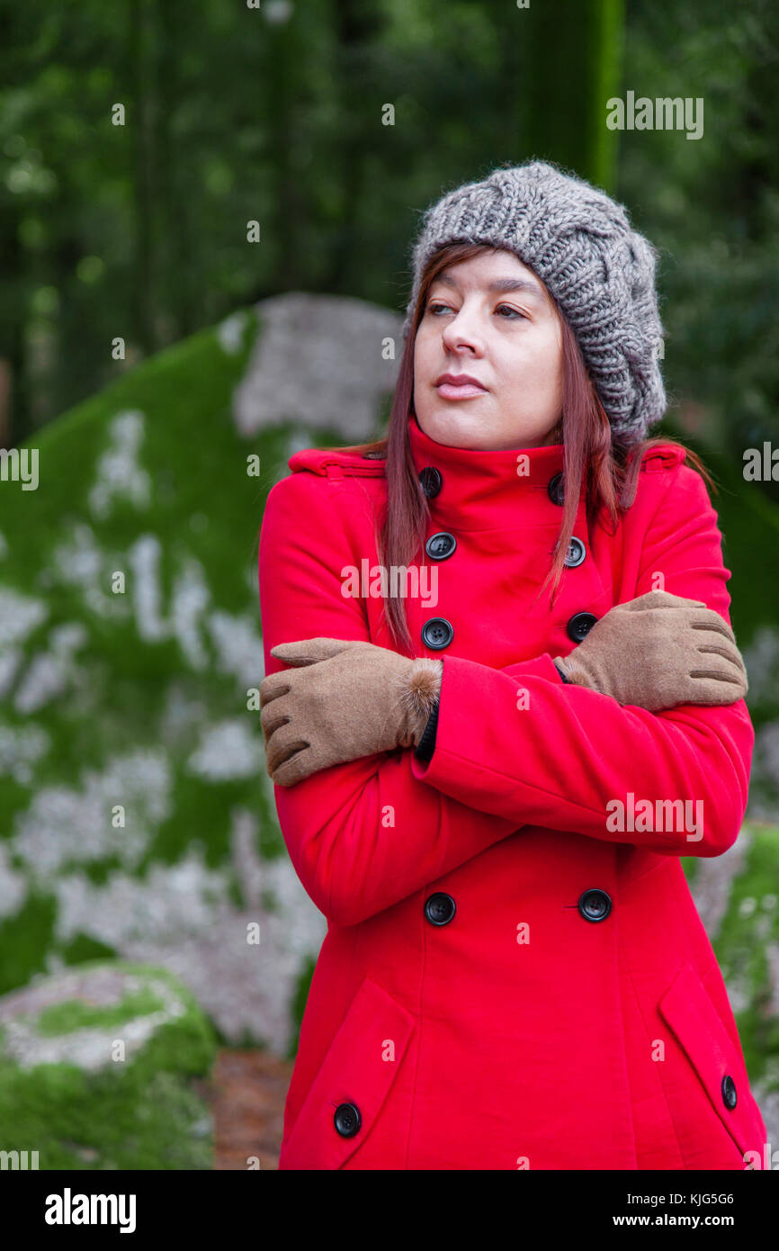 Young woman lost on a forest shivering with cold and embracing or holding herself, wearing a red long coat or overcoat, a beanie and gloves during fal Stock Photo