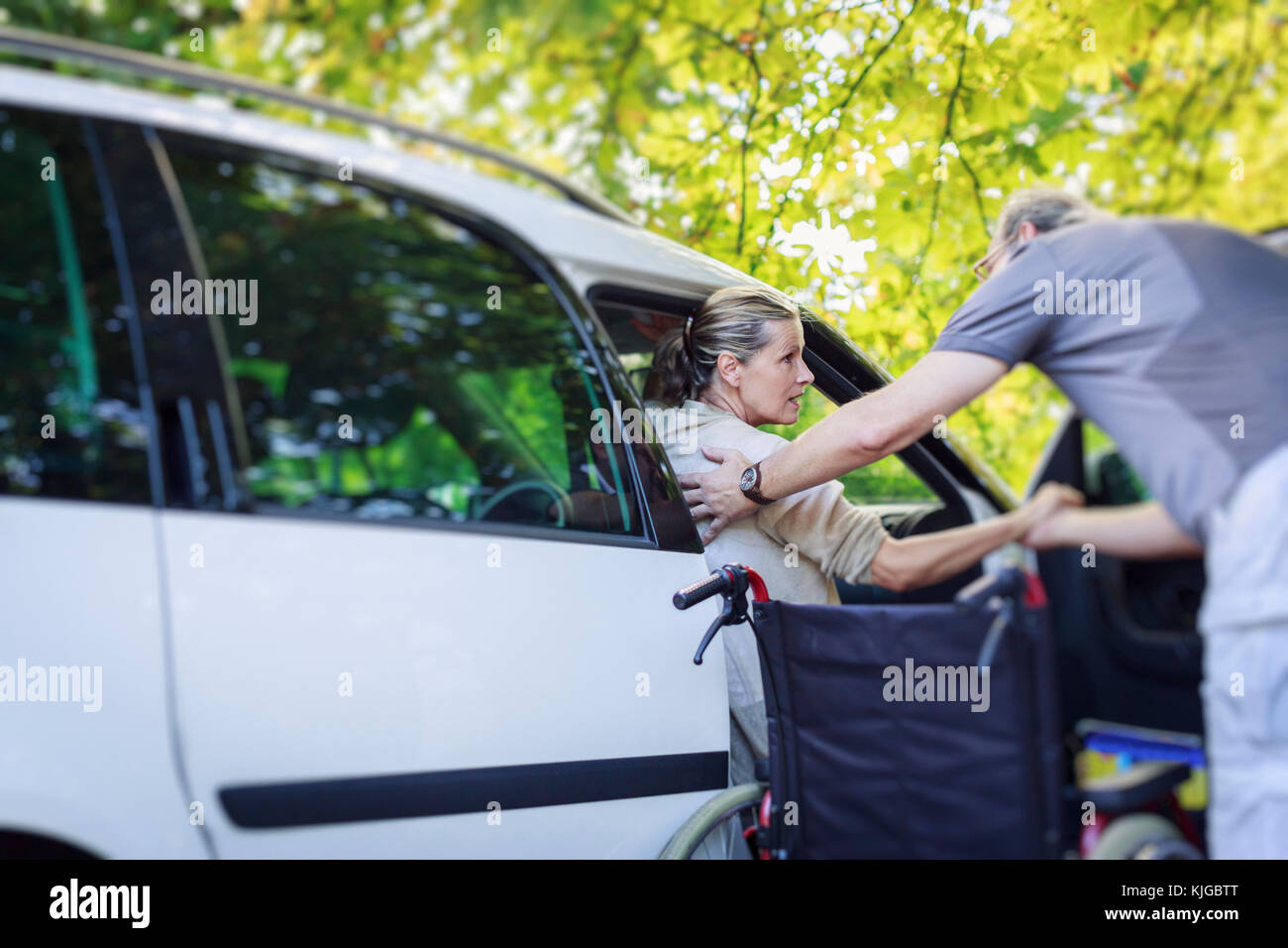 Husband helping his wife getting out of car next to wheelchair Stock