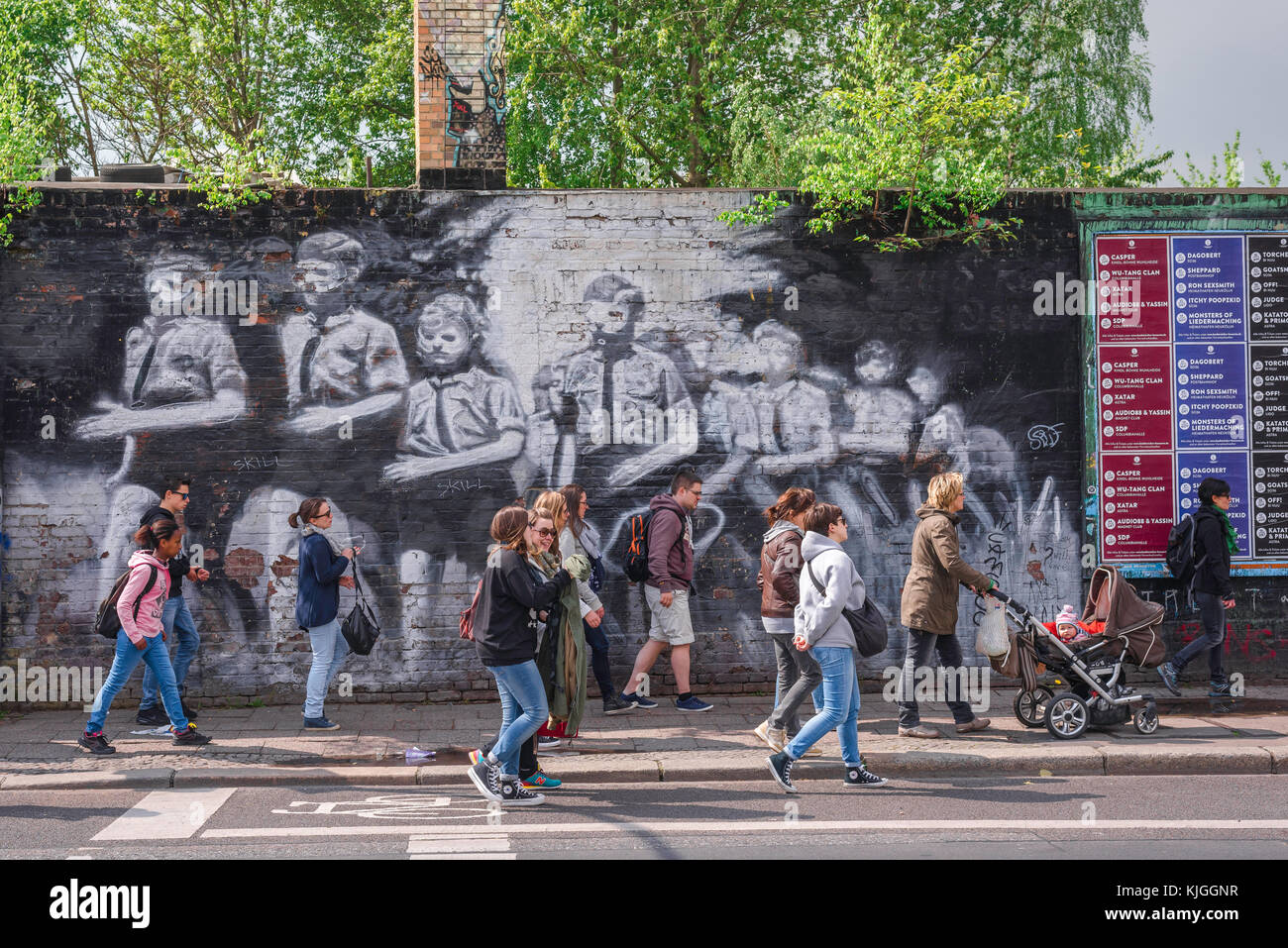 Berlin wall, people walk past a mural depicting Hitler Youth in stylised form painted on the Berlin Wall, East Side Gallery, Friedrichshain, Berlin. Stock Photo