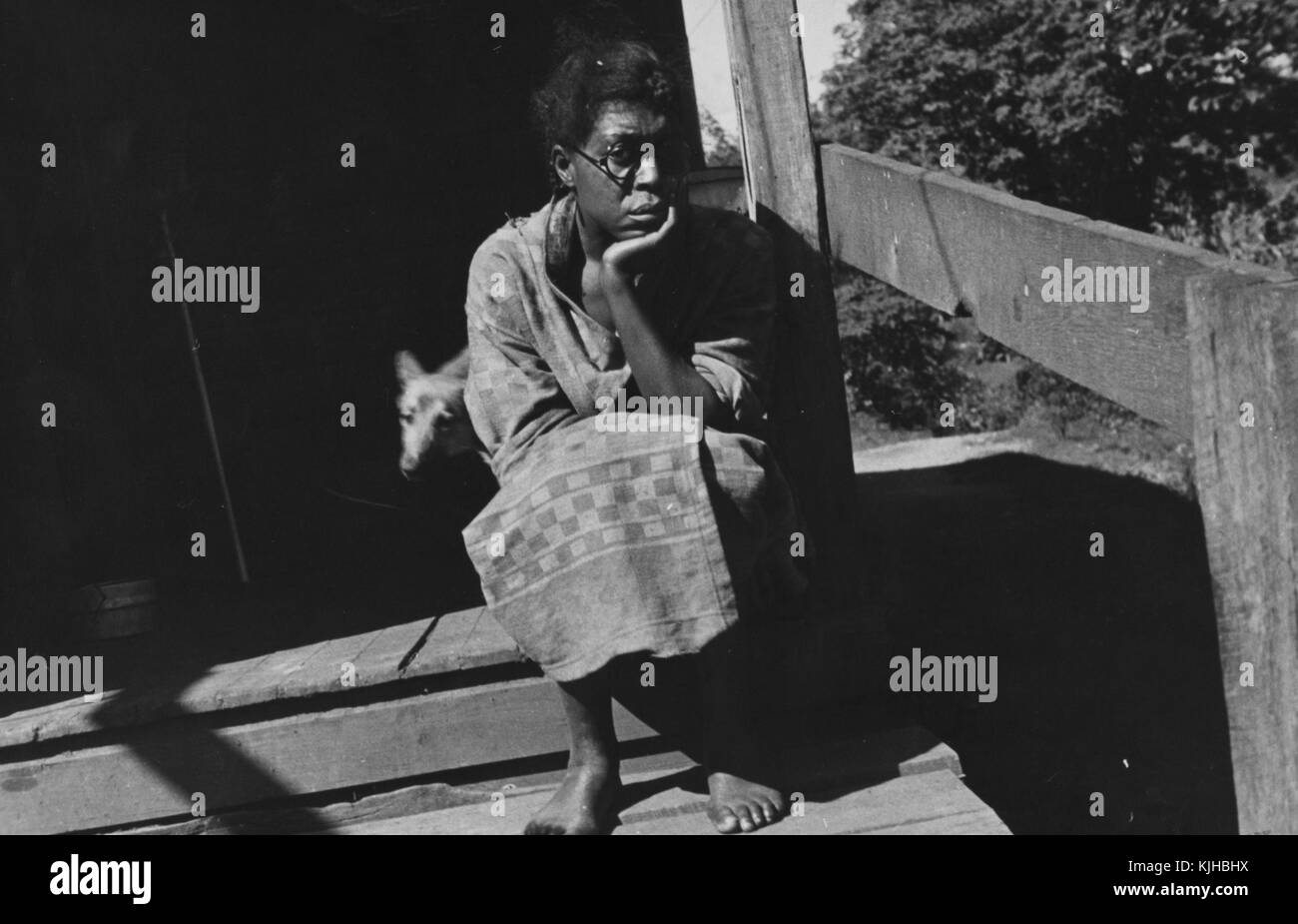 A photograph of a woman wearing a simple patterned dress, she is sitting on her wooden porch while she waits for a relief agent from the Farm Service Agency to meet with her, the FSA was working at the time to improve the lives of farmers around the country, her dog is coming out of the house behind her, Scott's Run, West Virginia, 1935. From the New York Public Library. Stock Photo