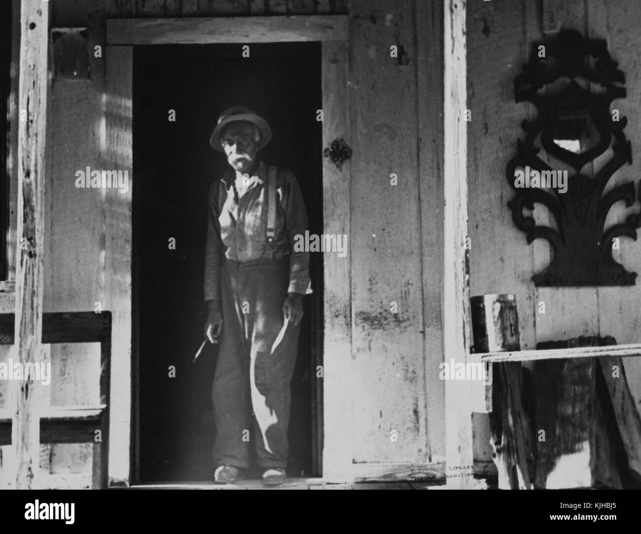 A photographic full length portrait of a sharecropper, he is wearing a button down shirt and pants held up by suspenders, he is standing in the doorway of his home, the paint is peeling from the porch of his home which contains a bench, a decorative mirror, lumber, and a can of paint, Arkansas, 1935. From the New York Public Library. Stock Photo