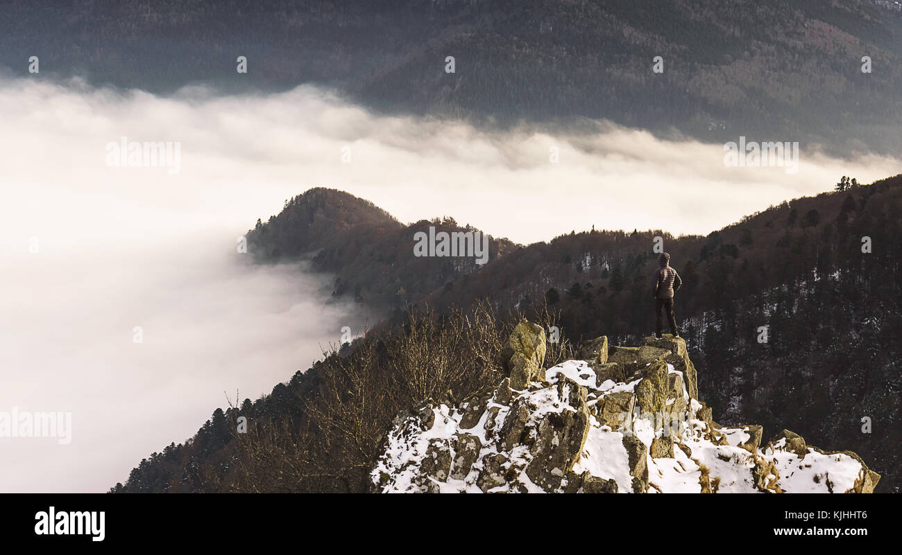A hiker standing on a snowy peak, looking at a cloud inversion in the mountains in early winter. Wanderlust in the Vosges (France). Stock Photo