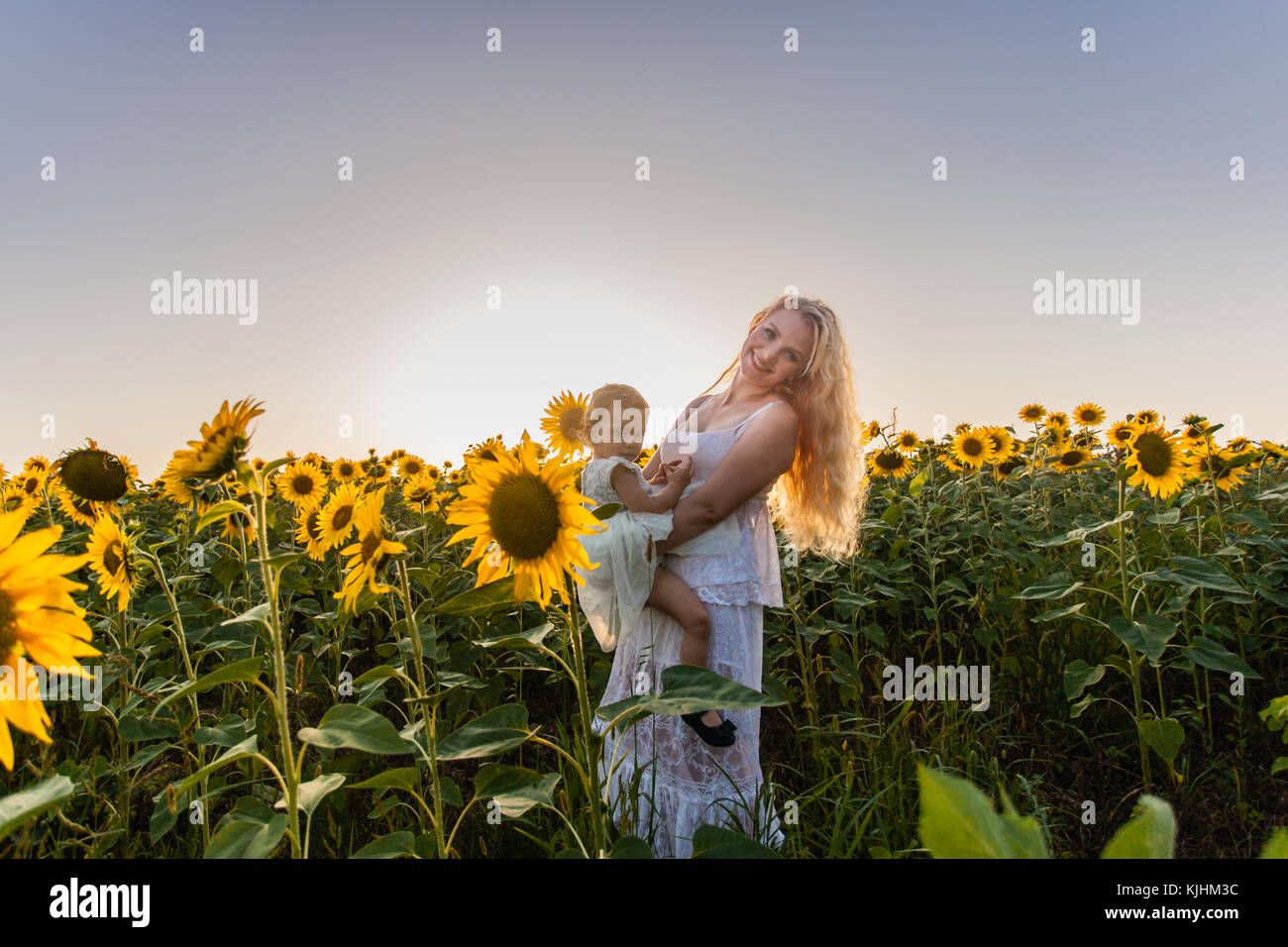 Happy mother hugs her daughter in a sunflower field, white clothes