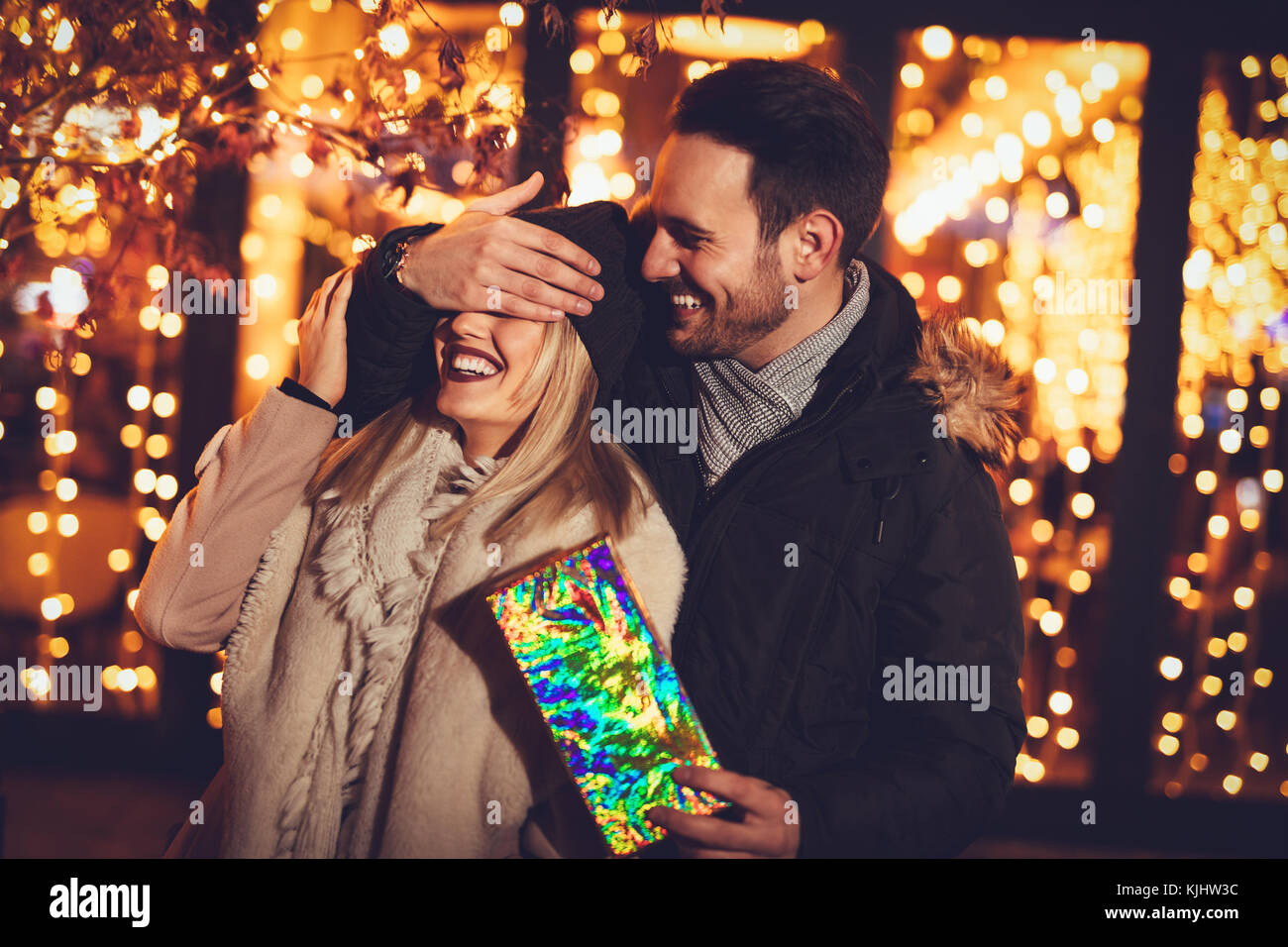 Beautiful cheerful couple celebrating Christmas in the city street Young man covering the eyes of happy surprised girlfriend and giving her present