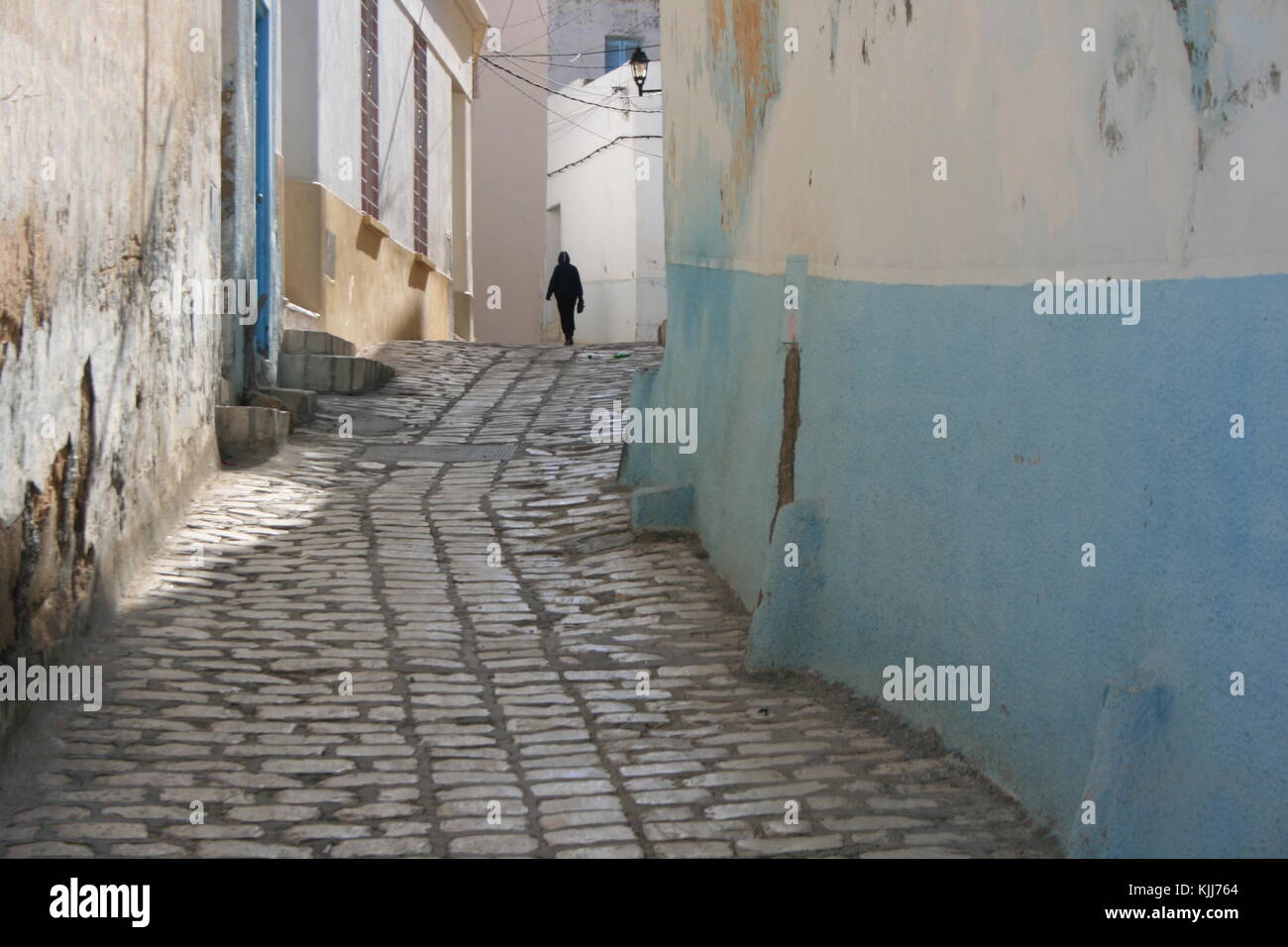 silhouette of a lonely woman walking at the end of a narrow old alley of hamammet in tunisia among colored white and blue walls Stock Photo