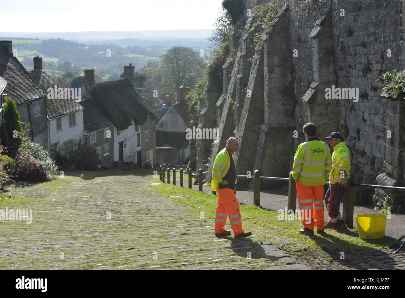 Three workmen discussing clearing the grass from the cobblestones of Shaftesbury Gold Hill, Dorset, England Stock Photo