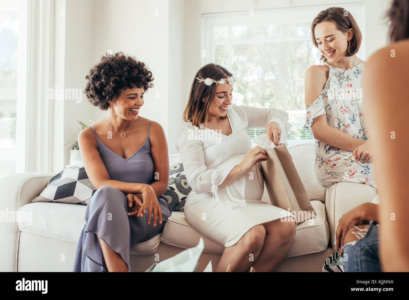 Happy pregnant woman with female friends at baby shower. Expecting woman sitting on sofa and opening gifts during her baby shower. Stock Photo