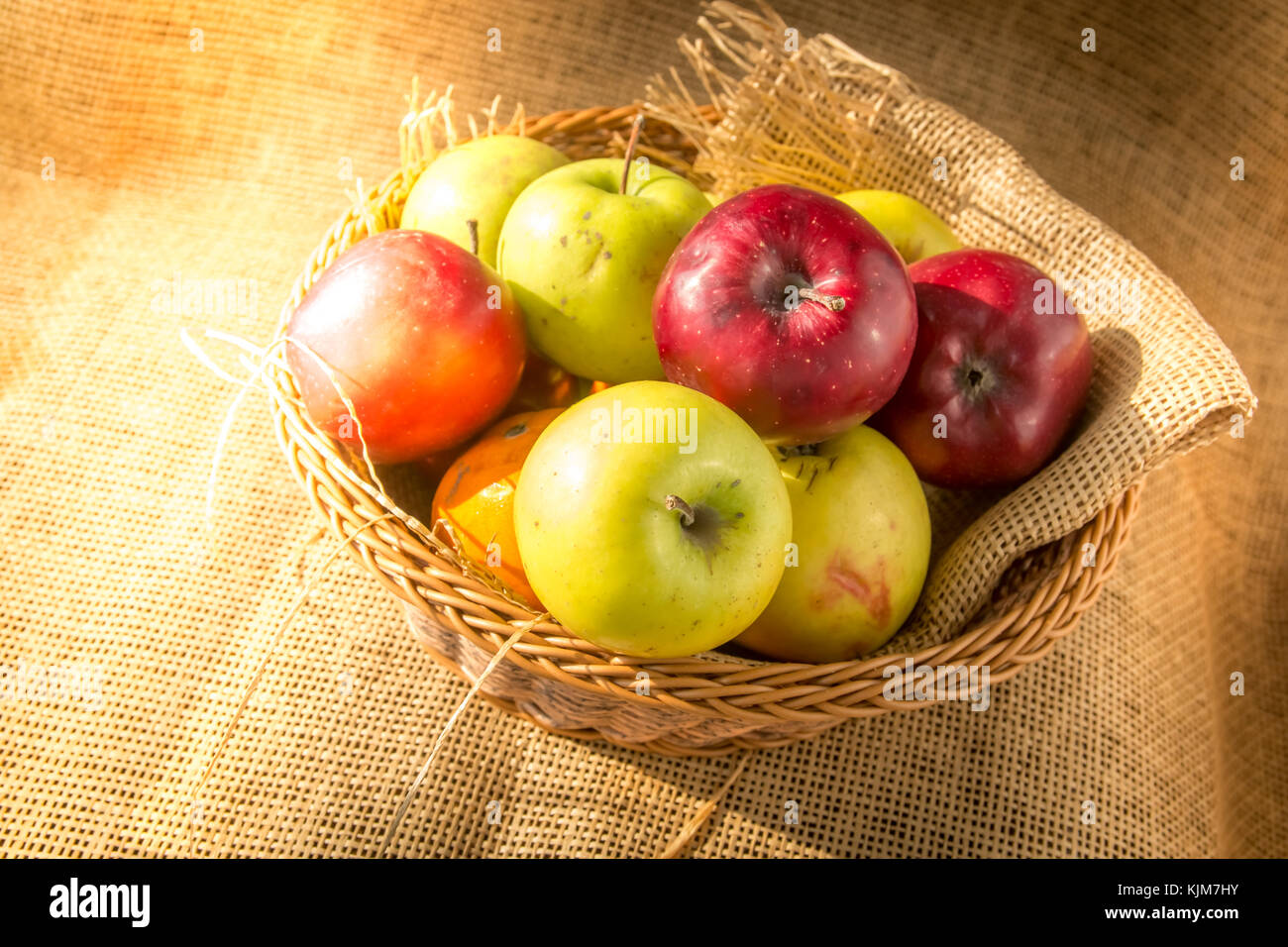 Green and red apples in a basket on burlap background with morning sun shine. Stock Photo